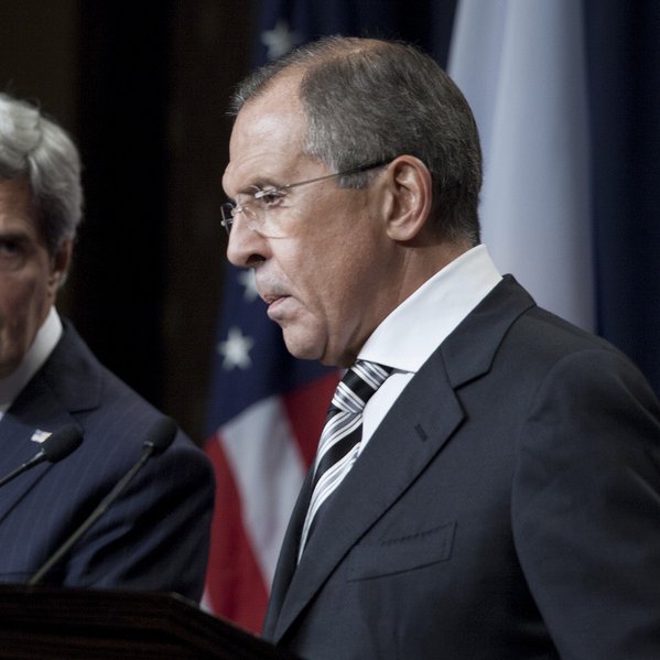 L'image montre deux hommes en tenue formelle, debout à un podium pendant une conférence de presse. L'un est à gauche, avec les cheveux gris et portant un costume bleu, et l'autre à droite, avec les cheveux foncés et portant un costume noir. Ils semblent être en train de discuter sérieusement, avec des drapeaux en arrière-plan, suggérant un contexte diplomatique ou de relations internationales.