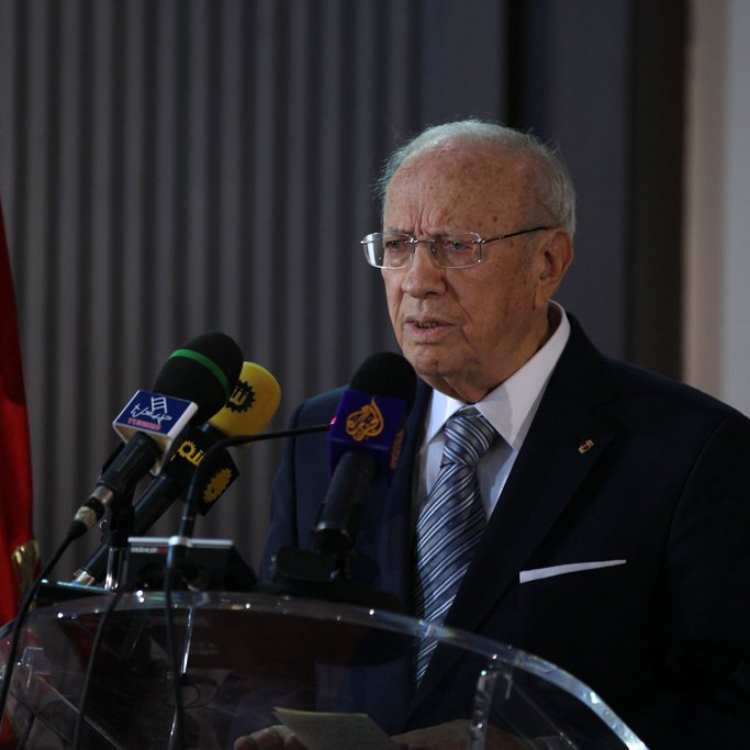 L'image montre un homme âgé qui se tient derrière un pupitre avec des micros. Il porte un costume sombre et une cravate, et semble s'adresser à un public. En arrière-plan, on peut voir un drapeau rouge, ce qui pourrait indiquer un contexte officiel, possiblement politique. L'atmosphère semble sérieuse et formelle.