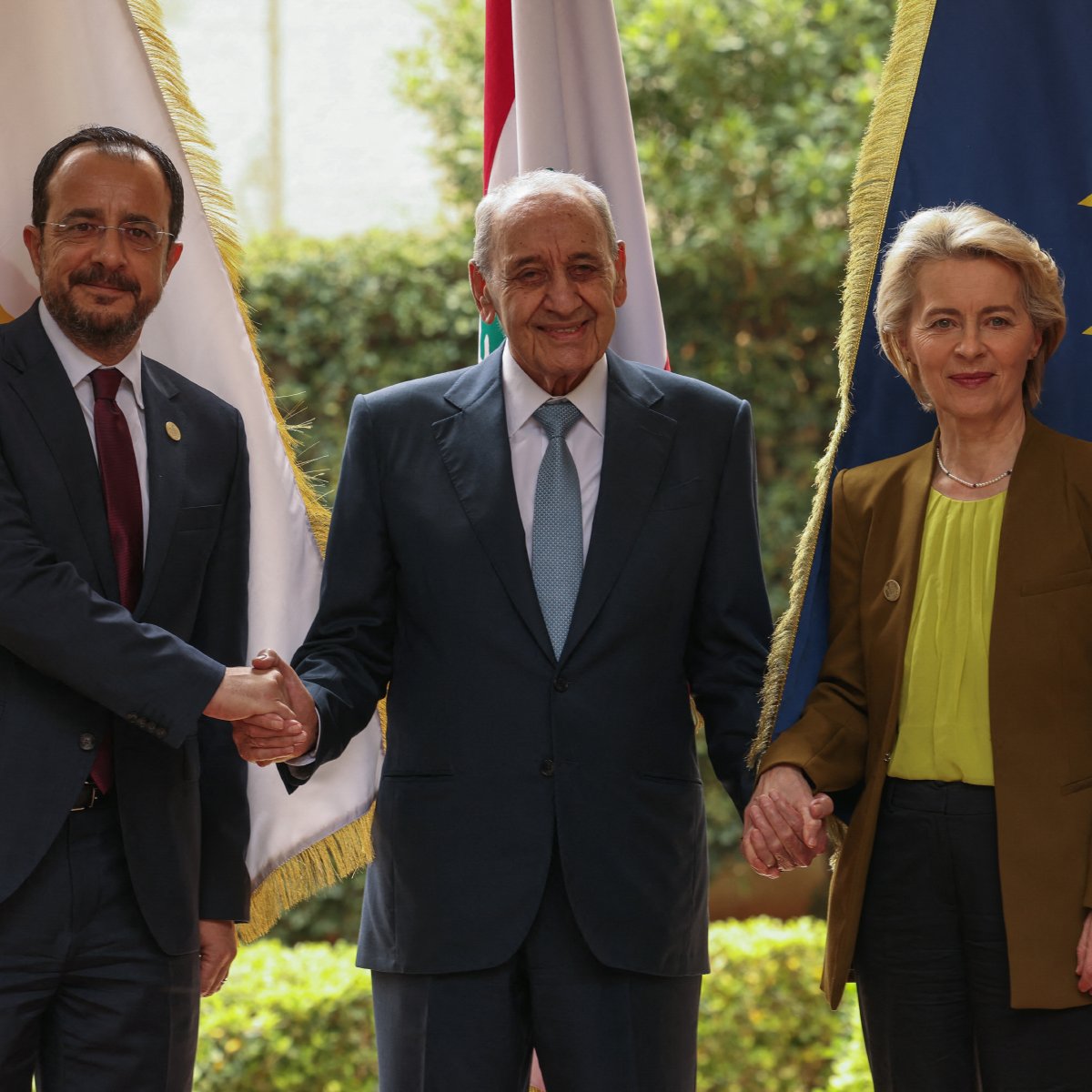 L'image montre trois personnes posant ensemble pour une photo. Au centre, un homme âgé souriant est entouré de deux personnes : à gauche, un homme en costume avec une cravate, et à droite, une femme en tenue professionnelle. Ils se tiennent la main en signe de coopération. En arrière-plan, on peut voir des drapeaux, dont un de l'Union européenne, ainsi que d'autres drapeaux nationaux. L'environnement est verdoyant et semble être en extérieur.