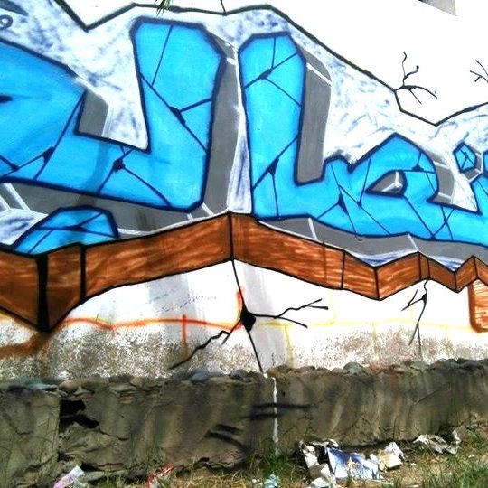 الصورة تظهر جدارية من فن الجرافيتي على حائط. الكلمة المكتوبة باللون الأزرق الفاتح تتوسط الجدارية، وتحيط بها بعض الخطوط والتفاصيل باللون الأسود. يبدو أن الجدارية تجذب الانتباه بلونها الزاهي وتصميمها الفني. الحائط يظهر بعض التشققات، مما يضيف إلى الجمالية الفنية للجدارية محاطة بالعشب والأوساخ في الأسفل.