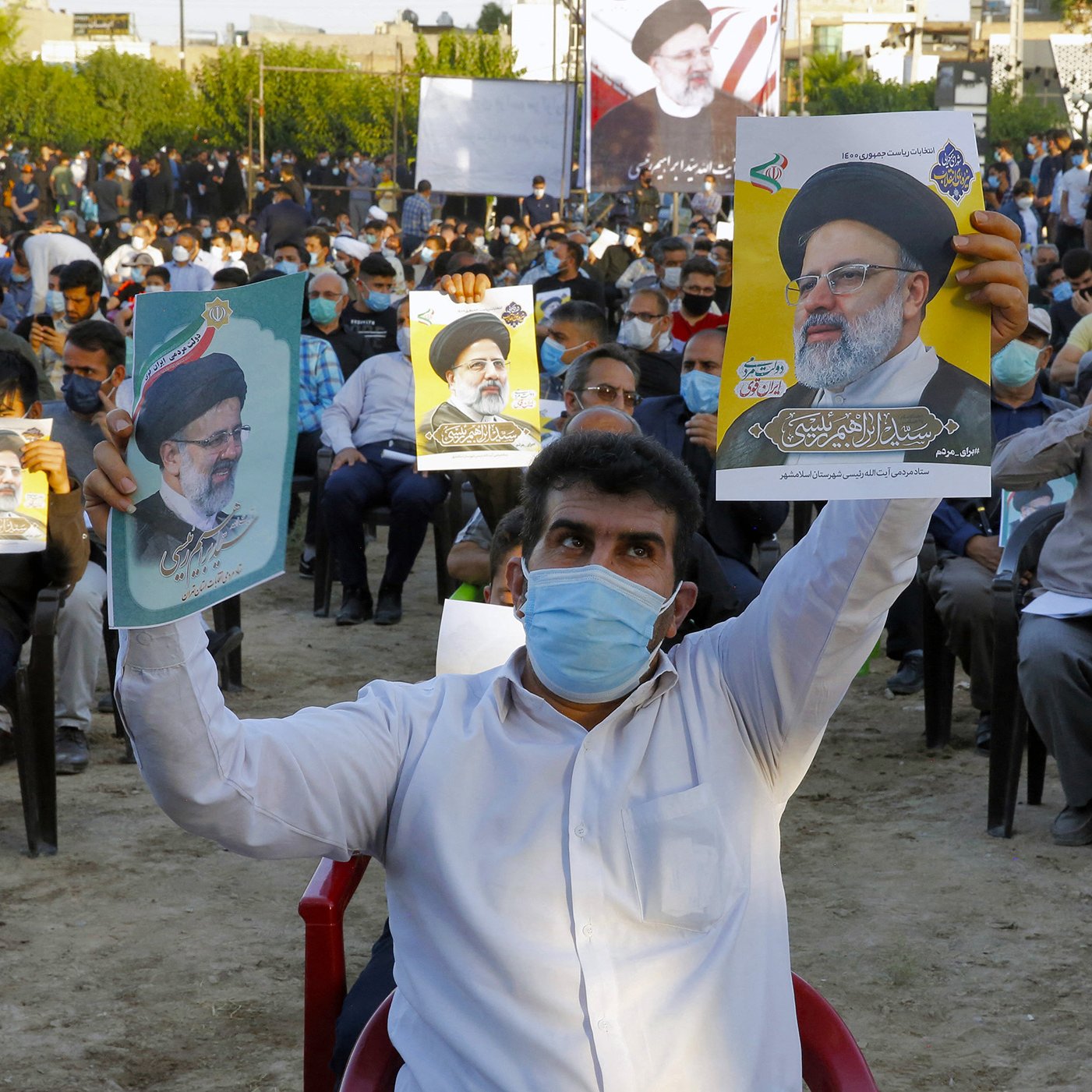 The image shows a large crowd of people seated outdoors, wearing masks. Many attendees are holding posters or pictures of a man, who appears to be a political figure, indicating support for him. The setting looks like a public gathering or rally, with trees in the background and banners visible. The mood appears to be one of engagement and solidarity among the participants.