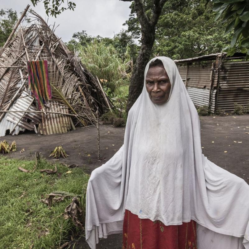 Sur cette image, on voit une femme debout au premier plan, portant un vêtement traditionnel avec un voile qui couvre sa tête. Son expression semble sérieuse ou réfléchie. À l'arrière-plan, on aperçoit une structure habitable, partiellement en bois et en matériaux naturels, qui semble être endommagée. La scène est entourée de verdure, avec des arbres et de l'herbe. L'ambiance générale évoque un contexte rural ou tribal, suggérant une connexion forte avec la nature et un mode de vie simple.