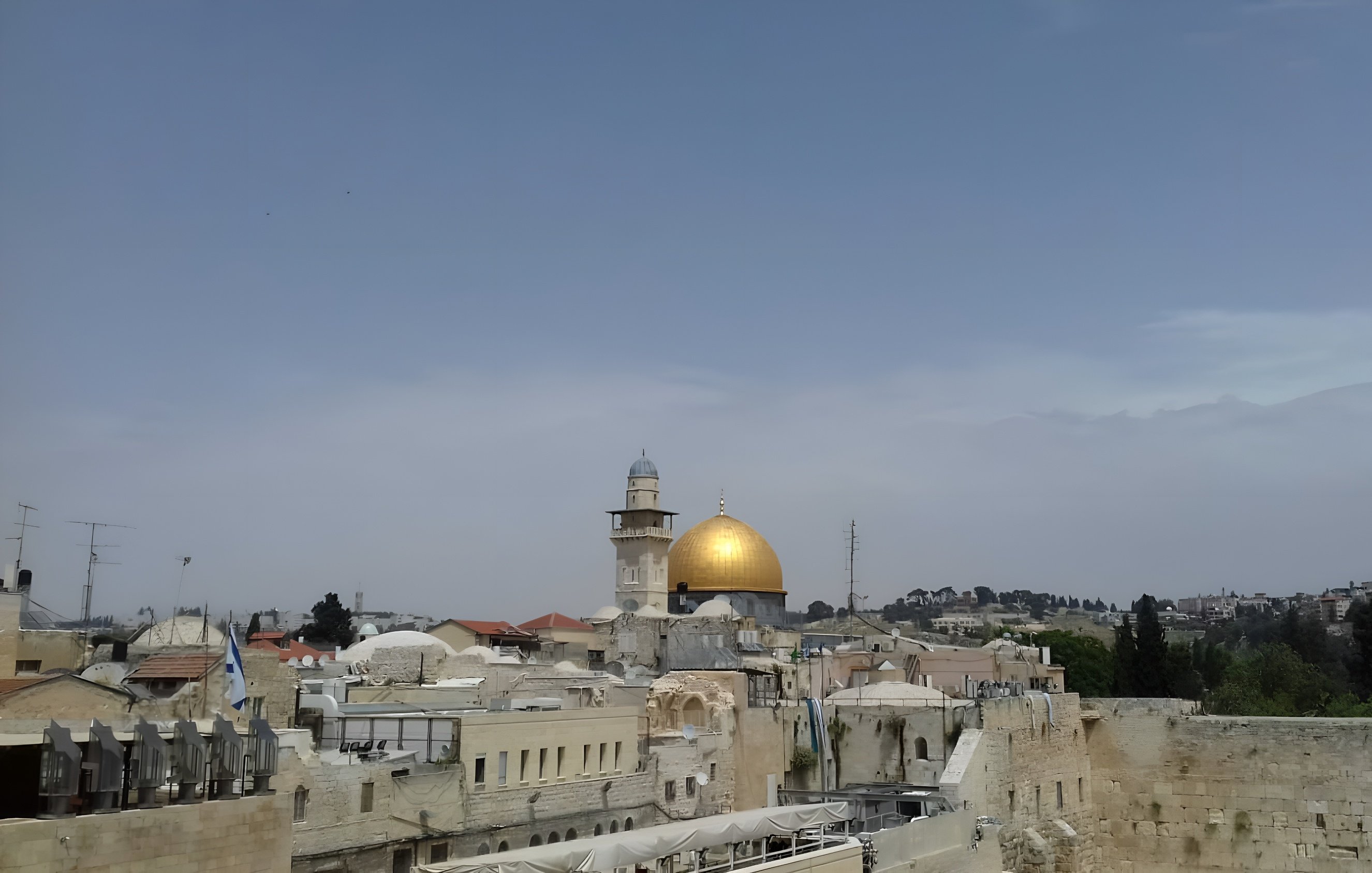 L'immagine mostra una vista panoramica di una città con una predominante cupola dorata, che è probabilmente il Dôme della Roccia, un noto sito religioso a Gerusalemme. Intorno ci sono edifici con tetti piatti e una torre, mentre il cielo appare sereno e leggermente nuvoloso. Gli alberi e le colline sullo sfondo aggiungono profondità alla scena.