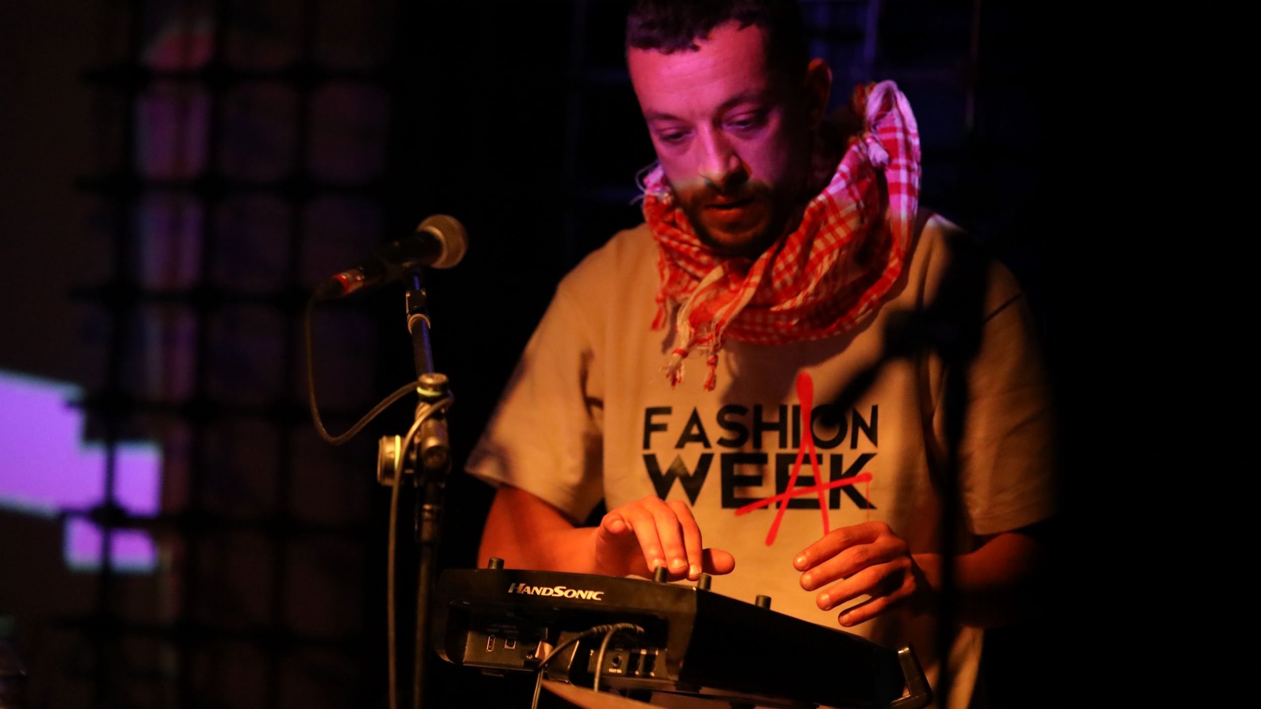 L'image montre un homme en train de performer sur scène. Il porte un t-shirt avec le mot "FASHION" écrit dessus et un foulard autour du cou. Il utilise un petit appareil électronique, probablement un synthétiseur ou un contrôleur musical. Le fond est légèrement éclairé avec des lumières colorées, créant une ambiance dynamique. Le microphone est également visible à côté de lui, indiquant qu'il pourrait chanter ou parler en même temps. L'atmosphère semble électrique et captivante.