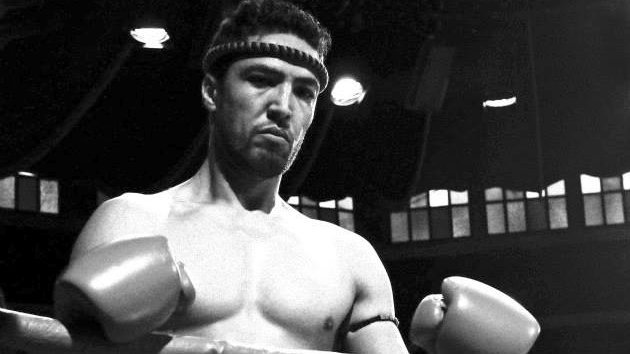 L'image montre un boxeur debout dans un ring. Il est torse nu et porte un bandeau autour de la tête. Ses poings sont protégés par des gants de boxe. Son expression semble concentrée et déterminée. Le style de l'image est en noir et blanc, ce qui lui donne une ambiance nostalgique et intemporelle. Le ring et l'arrière-plan suggèrent un environnement de compétition.