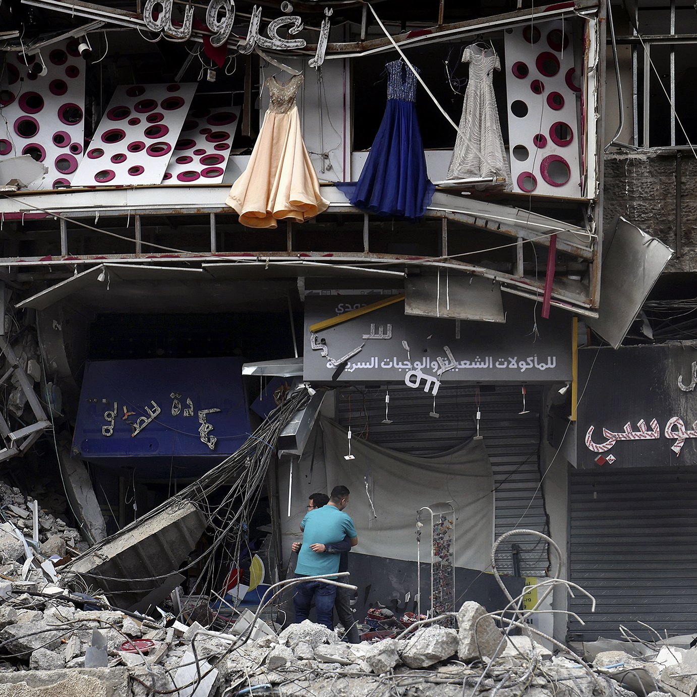 تظهر الصورة مشهدًا من منطقة دمار. هناك مبنى مهدوم جزئيًا يظهر فيه بعض الملابس المعلقة، مثل فساتين، بالإضافة إلى واجهات متاجر متضررة. يلاحظ في الأسفل شخصان يتفقدان المكان وسط الأنقاض. المشهد يعكس آثار تدمير كبير والخراب الذي لحق بالمنطقة.