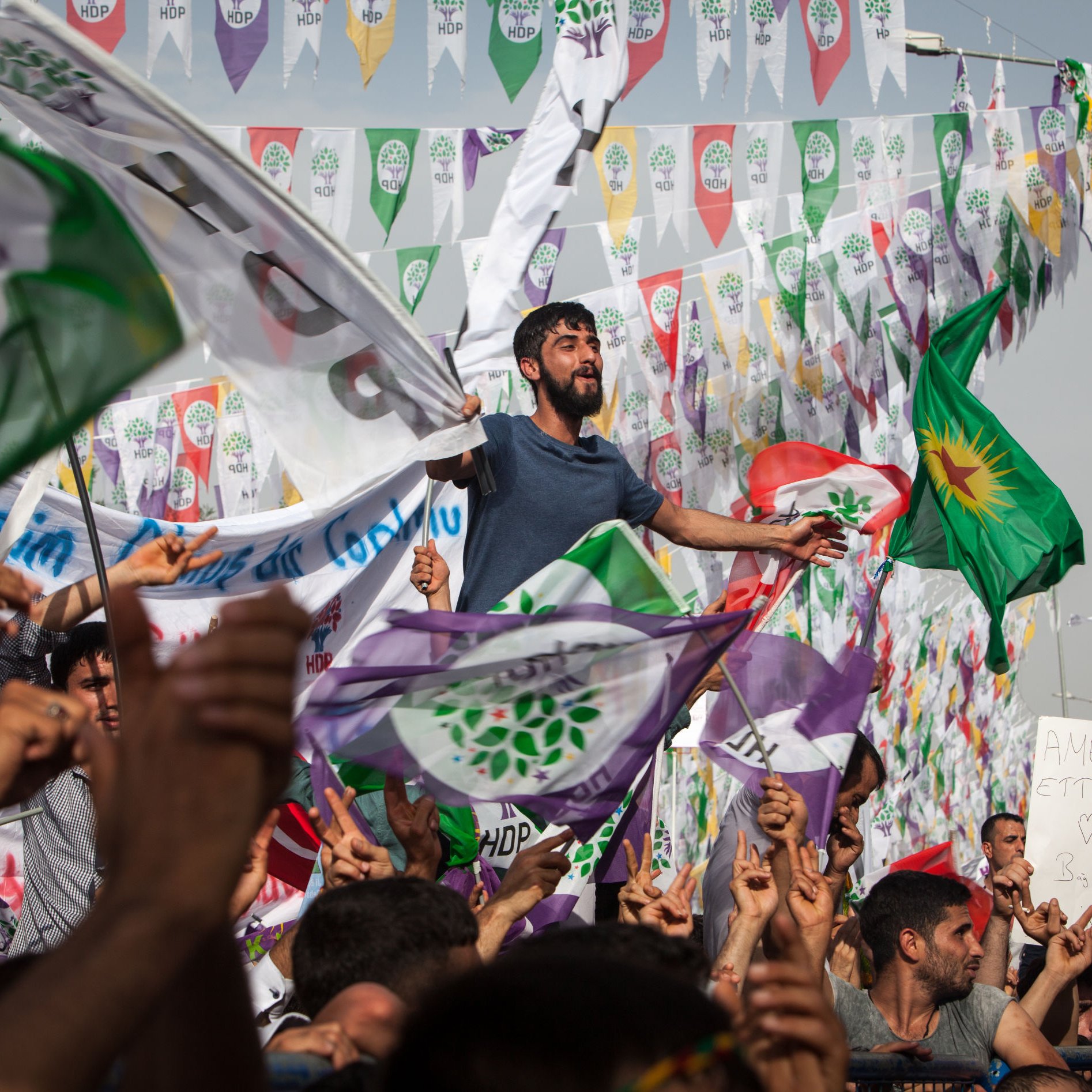 L'image montre une foule rassemblée lors d'un événement politique ou d'une manifestation. On peut voir des personnes brandissant des drapeaux, dont certains portent les couleurs et les symboles associés à un parti. L'ambiance semble festive et engagée, avec des individus visibles qui expriment leur enthousiasme, certains levant les bras ou chantant. En arrière-plan, des fanions colorés flottent, ajoutant à l'atmosphère vibrante de l'événement.
