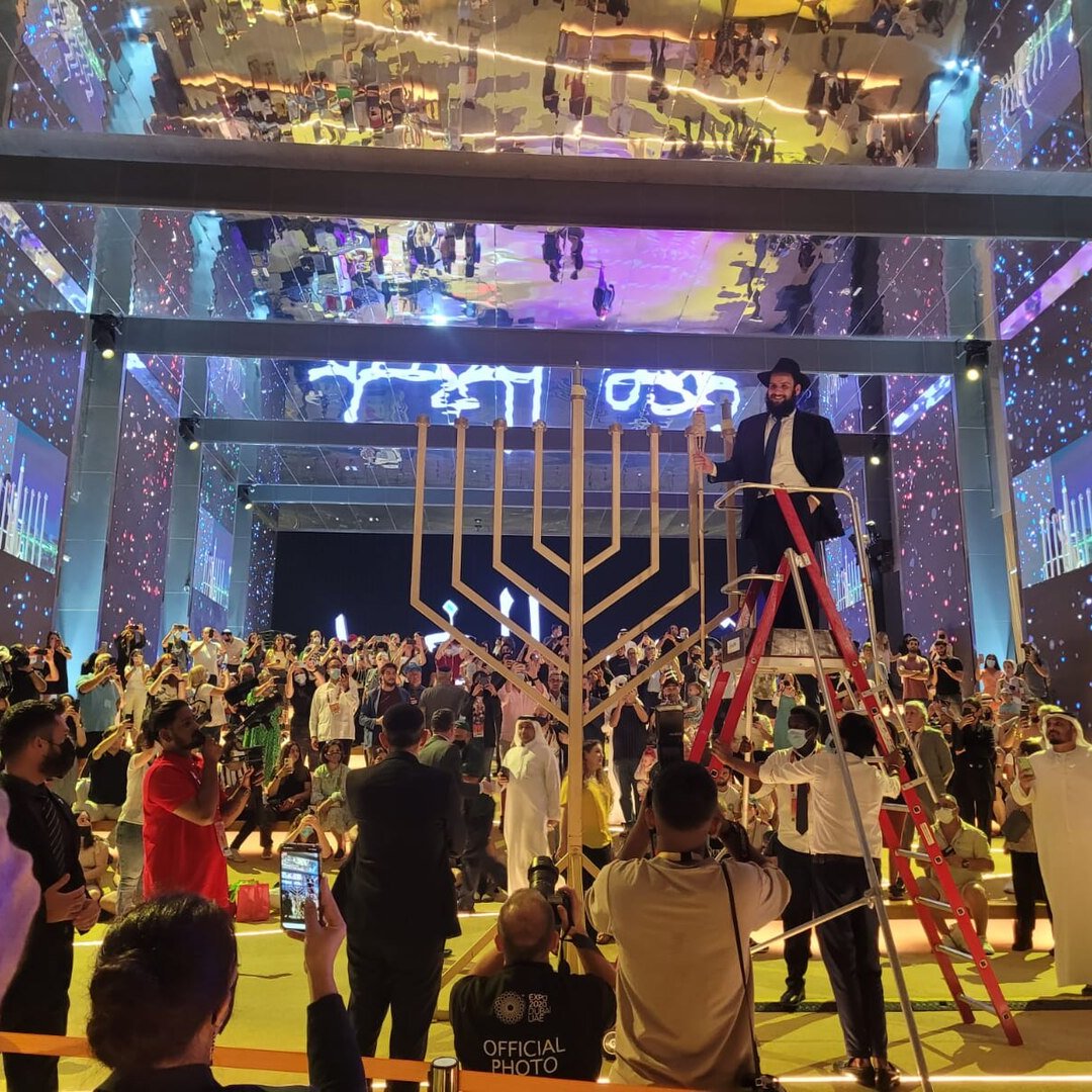 The image depicts a lively event taking place in a large, well-lit venue. In the center, there is a large menorah, which is a symbol associated with Hanukkah. A person is standing on a ladder, seemingly preparing to light the menorah. Surrounding the menorah, a diverse crowd is gathered, with some individuals clapping or taking photos. The background features colorful lights and projections, enhancing the festive atmosphere of the gathering. The overall scene reflects a sense of celebration and community.