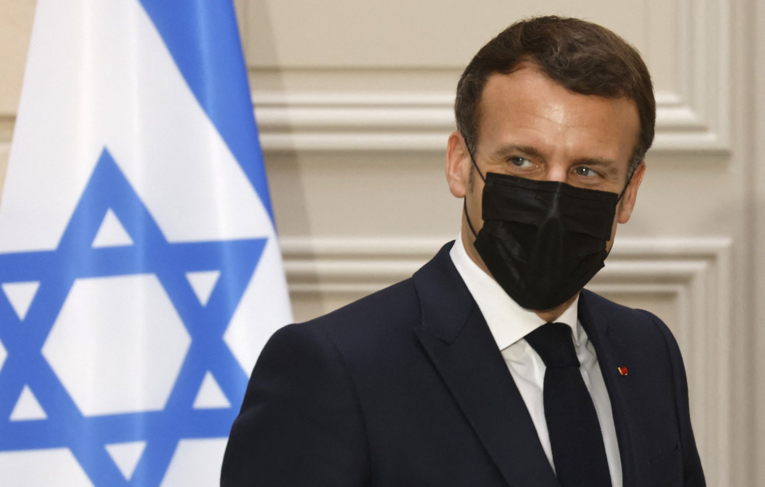 الصورة تظهر رجلًا يرتدي بدلة رسمية وقناعًا أسود. خلفه يوجد علم يحمل نجمة داود. يبدو الرجل في وضعية جانبية، حيث يلتفت نحو الجانب. تعكس الإضاءة في الصورة أجواء رسمية. لا يمكنني تحديد من هو هذا الشخص.