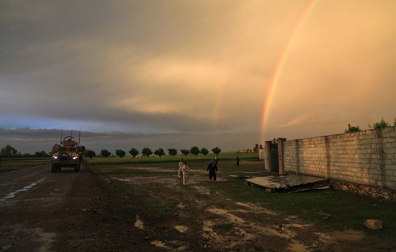 L'image montre un paysage rural sous un ciel dramatique, avec des nuages sombres et un ciel éclairci. On peut voir un arc-en-ciel double qui s'étend dans le ciel, ajoutant une touche de couleur. À gauche, un véhicule militaire est garé sur un chemin boueux, tandis qu'à droite, deux personnes marchent près d'un mur en béton. Le sol est humide, témoignant d'une récente pluie, et des arbres sont visibles en arrière-plan, soulignant une atmosphère paisible malgré la présence militaire.
