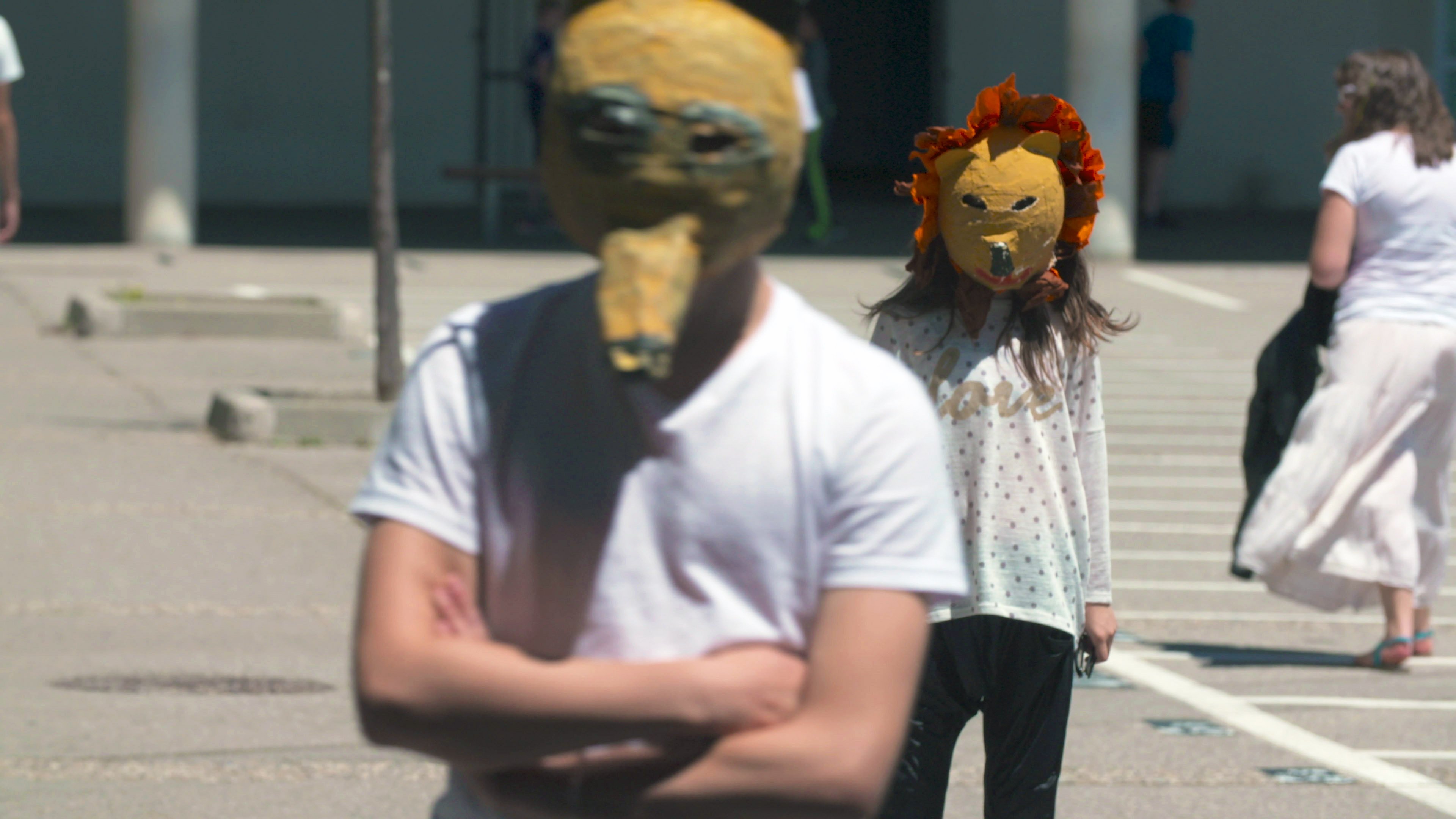 L'image montre deux personnes portant des masques originaux. L'un des masques a une forme de tête d'animal, tandis que l'autre est décoré de fleurs, évoquant un lion. Les personnes semblent se promener dans un espace extérieur, peut-être une place ou une cour. L'environnement est lumineux et rappelle une scène de performance artistique ou d'un événement communautaire.