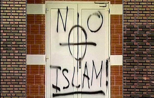 L'image montre une porte sur laquelle est peinte une inscription en spray noir. Le mot "NO" est écrit en haut à gauche, suivi d'un symbole ressemblant à une croix, et en dessous, le mot "ISLAM" est inscrit avec un point d'exclamation à la fin. Le fond de la porte et le mur environnant montrent des briques. L'ensemble de la scène évoque un message provocateur.