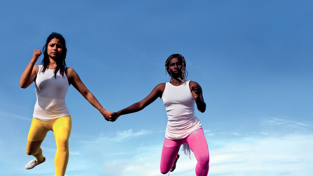 L'image montre deux femmes qui semblent flotter dans les airs, se tenant par la main. Elles portent des vêtements de sport, l'une en leggings jaunes et l'autre en leggings roses, avec des t-shirts blancs. En arrière-plan, on peut voir une ville urbaine s'étendre sous elles, avec un ciel bleu clair. Cette composition crée une sensation de liberté et de mouvement dynamique.
