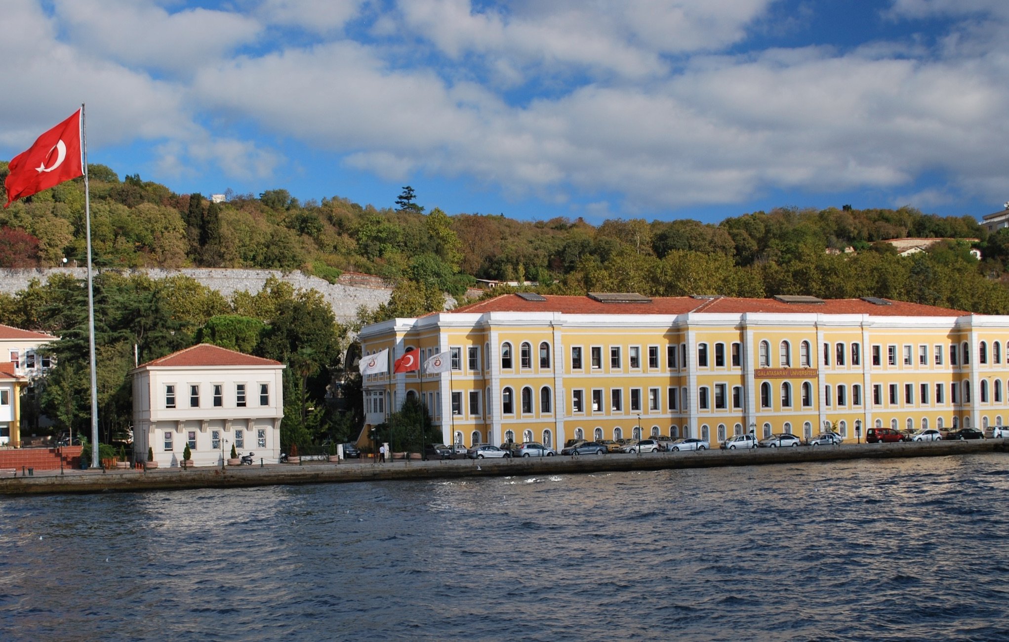 L'image montre un bâtiment jaune surplombant une étendue d'eau, probablement un bras de mer. À gauche, un grand drapeau turc flotte fièrement. On aperçoit également des arbres verdoyants en arrière-plan, ainsi qu'un ciel partiellement nuageux. Le bâtiment semble être de style architectural ancien, avec des fenêtres à volets et un toit en tuiles. Il y a des voitures garées le long de la rue en face du bâtiment. L'ensemble dégage une atmosphère paisible et pittoresque.