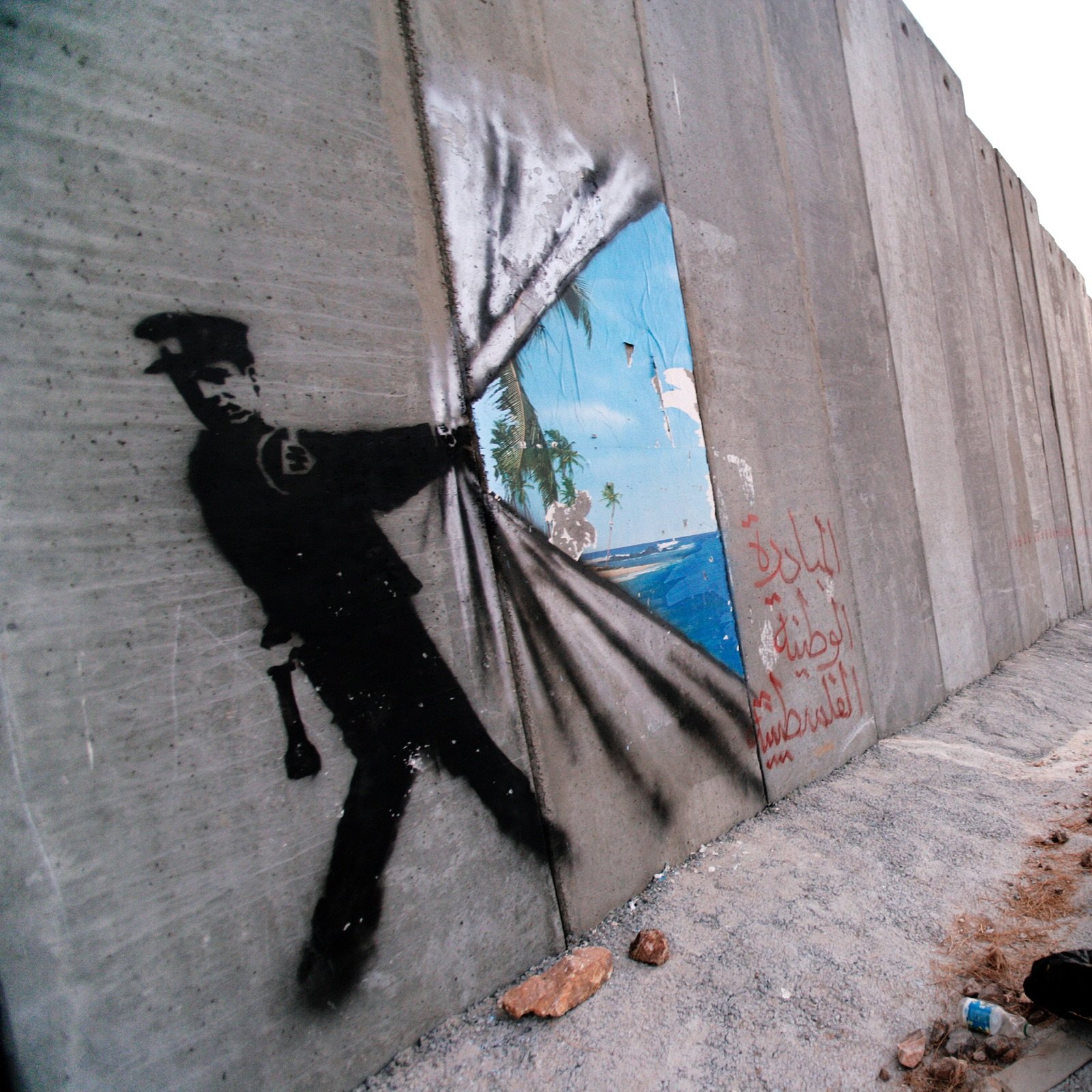 L'image montre un mur de béton, probablement un mur de séparation ou de sécurité, sur lequel est peint un graffiti représentant un policier. Ce dernier semble tirer un rideau pour révéler un paysage idyllique, avec des palmiers et un ciel bleu, symbolisant un contraste entre la réalité de l'environnement urbain et un espace rêvé ou libre. Sur le mur, on peut également voir des inscriptions en arabe. L'ensemble de l'image peut évoquer des thèmes de restriction, d'espoir et de liberté.