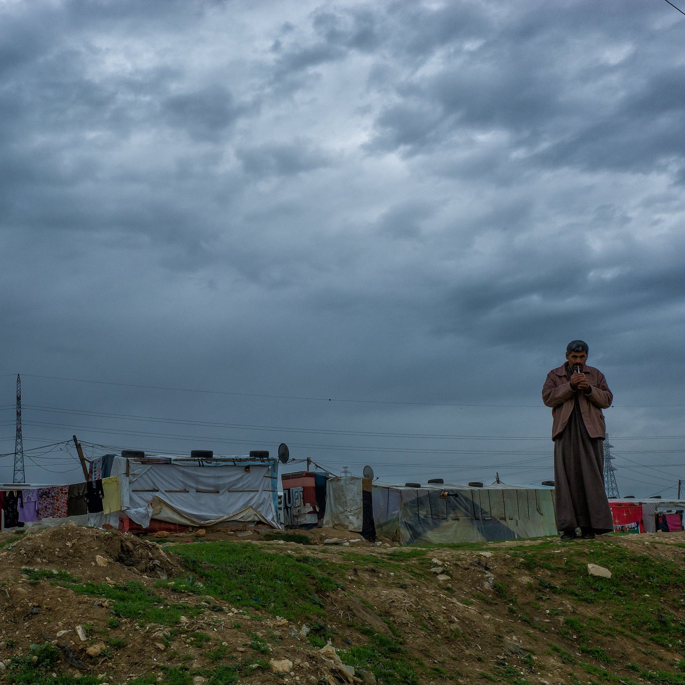 L'image montre une scène en extérieur sous un ciel nuageux. À droite, un homme se tient debout, apparemment pensif, avec une main près de son visage. En arrière-plan, on voit une rangée de tentes ou abris, probablement dans un camp, avec des murs en toile de différentes couleurs. Le sol est herbeux et légèrement surélevé. L'atmosphère est sombre en raison des nuages menaçants qui couvrent le ciel.
