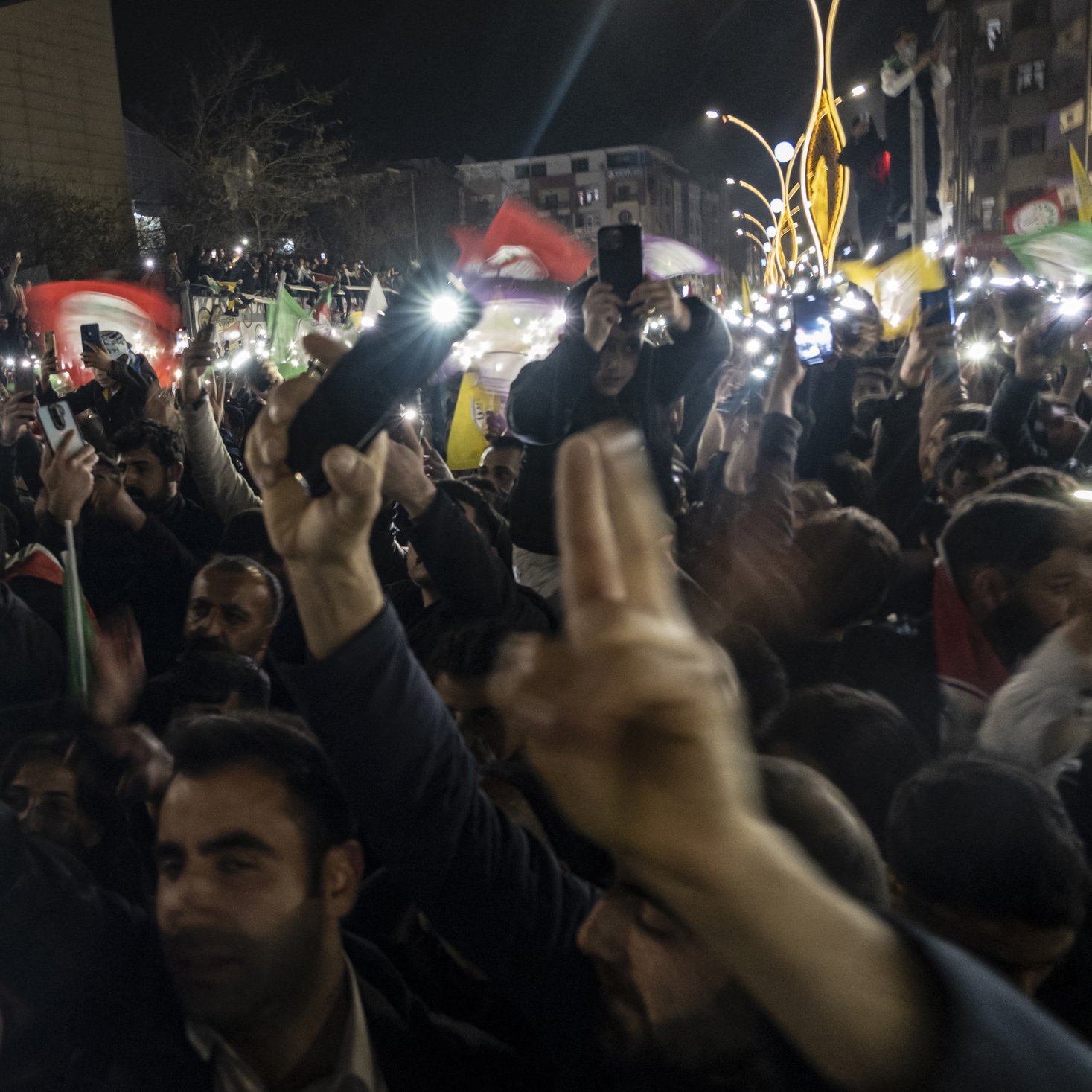 L'image montre une grande foule rassemblée dans un lieu public, probablement lors d'une manifestation ou d'une célébration nocturne. Les participants brandissent des drapeaux colorés et lèvent les mains, certains tenant des téléphones allumés qui éclairent la scène. L'atmosphère semble festive et énergique, avec des expressions de joie et d'engagement visible sur les visages des gens. Les bâtiments environnants sont sombres, accentuant les lumières des téléphones et des drapeaux.