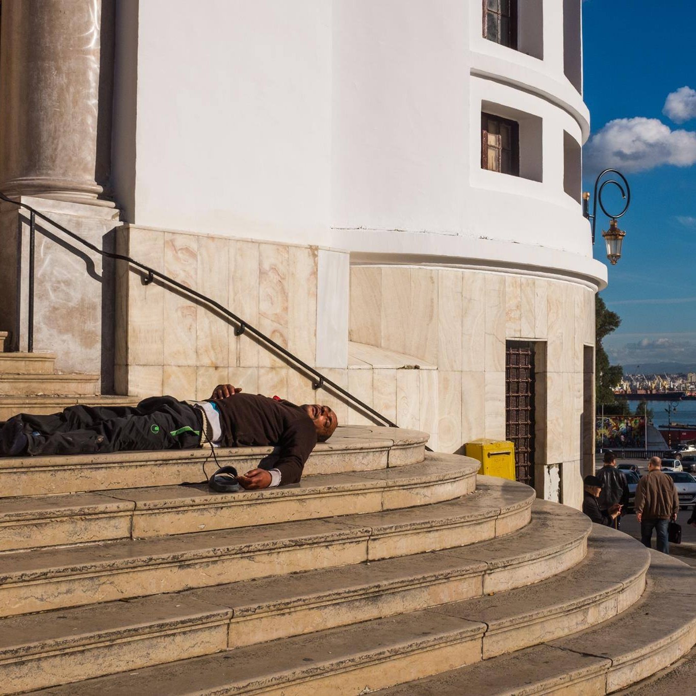 L'image montre un homme allongé sur les marches d'un bâtiment au style architectural classique, probablement en train de dormir ou en état de détresse. Le bâtiment est bien éclairé et ses murs sont de couleur claire, avec des colonnes visibles. En arrière-plan, on peut apercevoir une ville animée, avec des gens qui marchent et un port. Le ciel est bleu avec quelques nuages, ce qui donne une ambiance contrastée à la scène.
