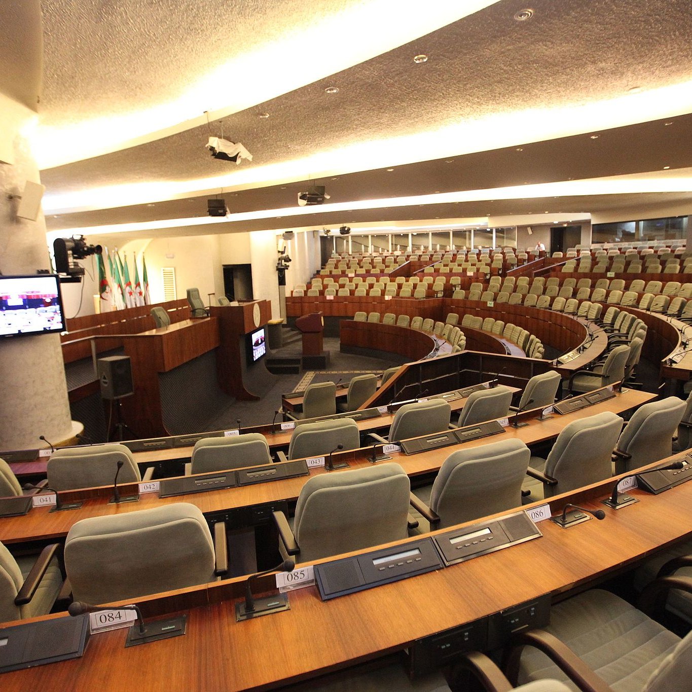 L'image montre une salle de réunion ou de débat, probablement une chambre législative. On y voit plusieurs rangées de sièges grisés disposés en arc de cercle autour d'une scène centrale. À l'avant, un bureau ou une tribune est visible, avec des équipements audiovisuels, comme un écran et des micros intégrés dans les tables. L'éclairage est doux et moderne, créant une atmosphère propice aux discussions. Les murs sont revêtus de matériaux contemporains, donnant une impression de sérieux et de fonctionnalité.