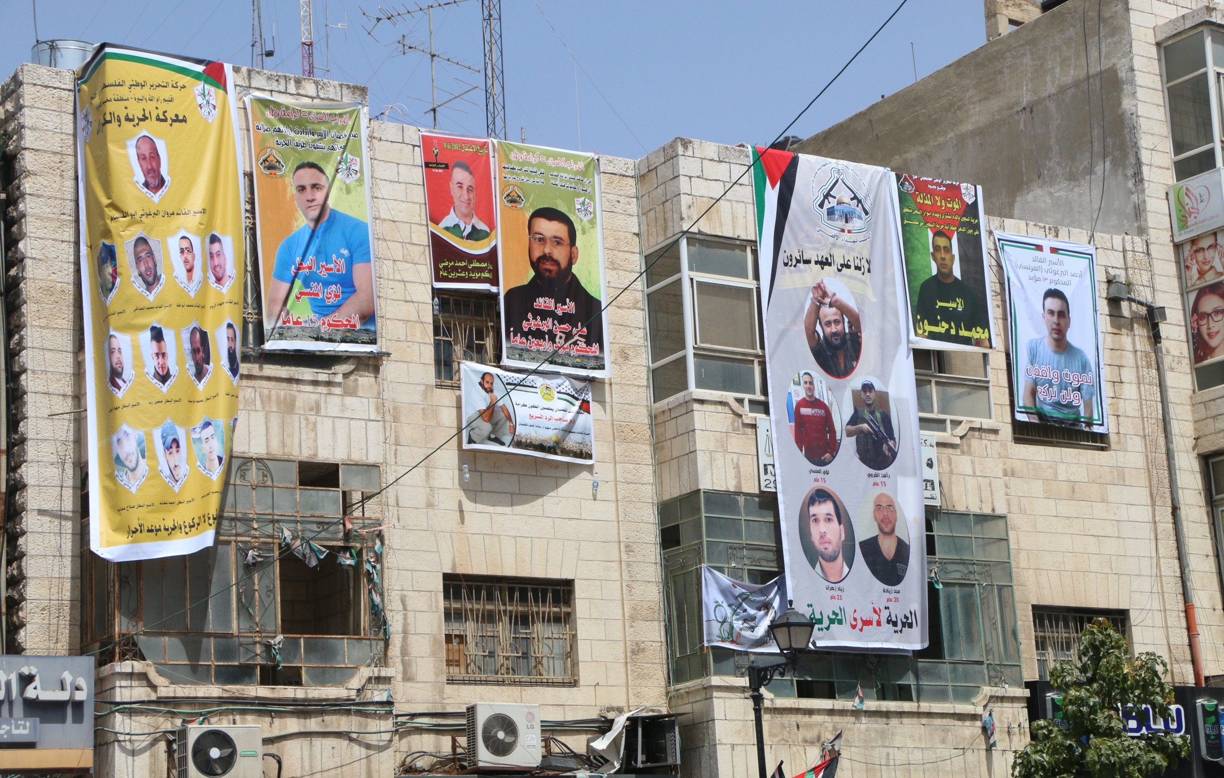 L'image montre un bâtiment avec plusieurs affiches et bannières accrochées sur sa façade. Ces affiches présentent des portraits de différentes personnes, probablement des figures politiques ou des militants, accompagnées de textes en arabe. Les couleurs des bannières sont vives, avec du jaune, du vert et du rouge, et beaucoup d'entre elles semblent commémorer ou soutenir des individus spécifiques. On peut également voir des drapeaux et d'autres éléments décoratifs liés à des mouvements politiques. L'environnement urbain donne une impression d'animation et de vie locale.