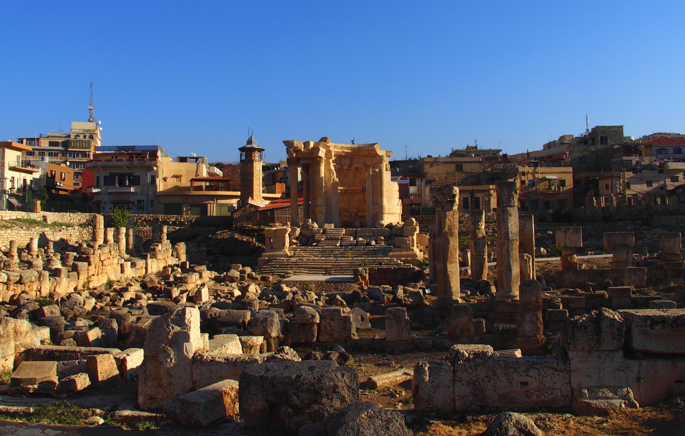 L'image montre un site archéologique avec des ruines anciennes, probablement d'un temple, entourées de colonnes et de blocs de pierre. En arrière-plan, on voit des bâtiments modernes et des structures urbaines s'élevant sous un ciel bleu. La lumière du soleil éclaire les ruines, créant un contraste entre l'ancien et le moderne. Ce lieu évoque une riche histoire et une importance culturelle.