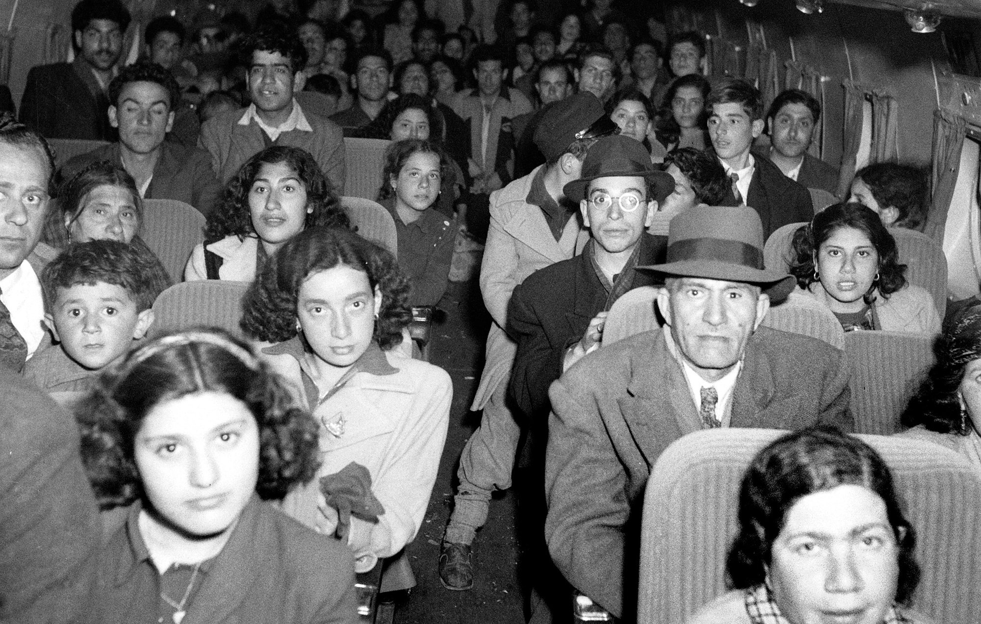 The image depicts a crowded interior of an airplane filled with passengers. There are men, women, and children seated in rows, looking directly at the camera. The individuals exhibit a mix of expressions, ranging from serious to curious. Many of them are dressed in clothing typical of earlier decades, suggesting that the photo may be historical. The cabin has a vintage appearance, and the layout and design of the seats reflect an earlier era of air travel.