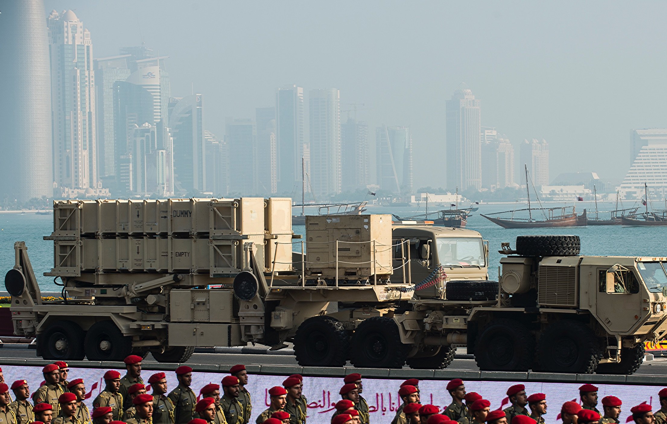 L'image montre des équipements militaires, probablement des systèmes de défense aérienne, positionnés sur des camions. En arrière-plan, on aperçoit une ville moderne avec des gratte-ciels et des bâtiments contemporains, ainsi qu'une étendue d'eau et des bateaux traditionnels. Au premier plan, des soldats en uniforme, peut-être d'une force armée, sont alignés, ajoutant une dimension cérémonielle à la scène. L'atmosphère semble être celle d'un défilé militaire ou d'un événement similaire.