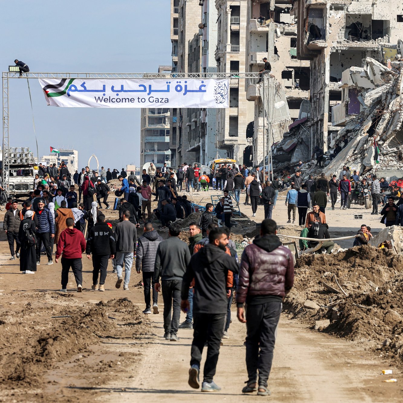 L'image montre une scène dans une ville en reconstruction, probablement à Gaza. On voit des bâtiments partiellement détruits, avec des débris éparpillés. De nombreuses personnes marchent dans la rue, certaines portant des gilets de bénévoles ou de secours. Un panneau en arabe et en anglais indique "Bienvenue à Gaza". L'ambiance dégage un mélange d'espoir et de désolation, avec des gens qui semblent se diriger vers un lieu particulier. Les conditions environnantes sont poussiéreuses et témoignent des récents conflits.