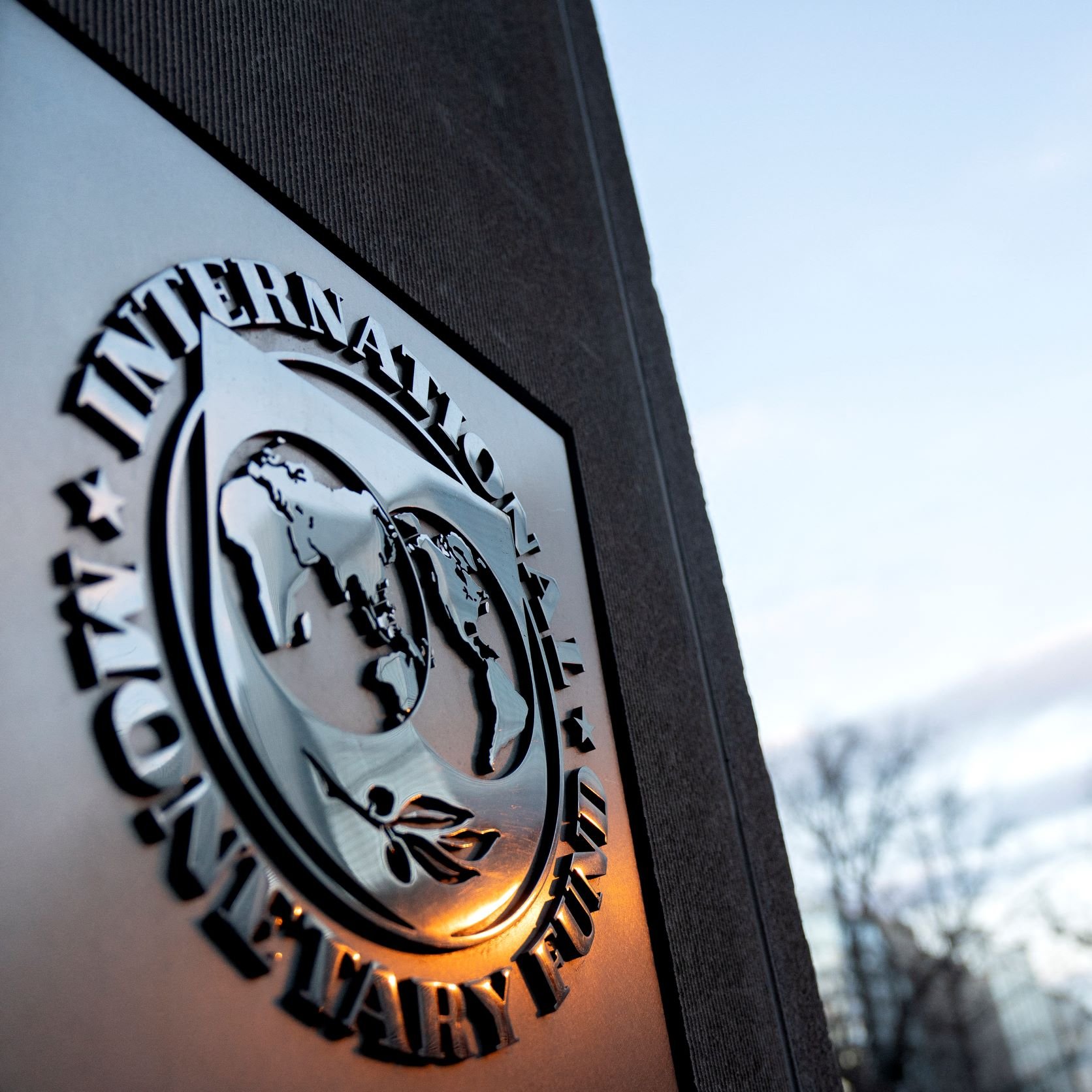 تصویر نشان‌دهنده لوگوی صندوق بین‌المللی پول (IMF) است که به‌صورت سه‌بعدی روی سطحی قرار دارد. در پس‌زمینه، ساختمان‌هایی با طراحی مدرن و آسمان روشن قابل مشاهده است. لوگو شامل نقشه‌ای از جهان و نمادهایی است که به فعالیت‌های اقتصادی و مالی اشاره دارند. این تصویر حس قدرت و اعتبار این نهاد بین‌المللی را منتقل می‌کند.