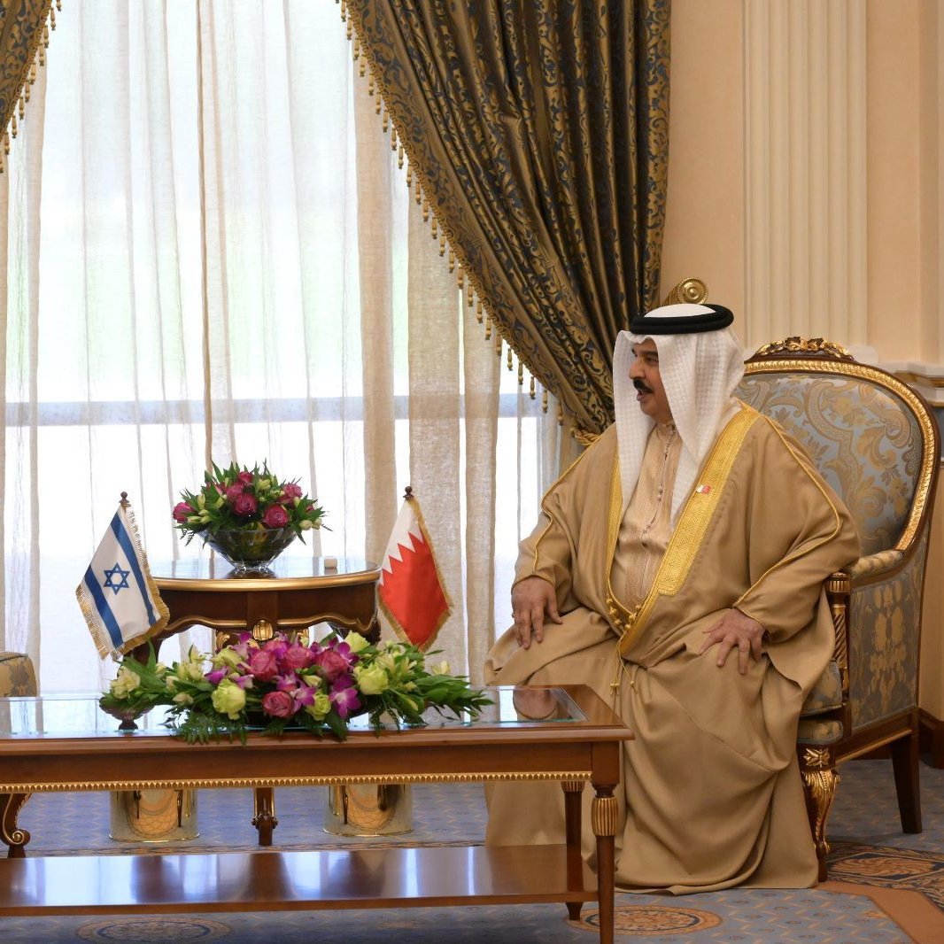 Two men in formal attire sit across from each other in a richly decorated room, discussing.