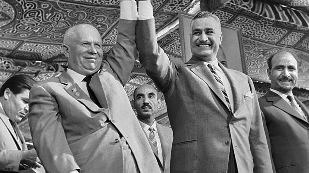 تظهر في الصورة مشهد تاريخي يجمع بين شخصين بارزين، حيث يحتفلان برفع أيديهما معًا تعبيرًا عن التضامن. الخلفية تبرز تفاصيل مميزة تشير إلى بيئة احتفالية، مع وجود عدد من الرجال الآخرين في الخلفية. يعكس هذا الموقف روح التعاون بين الشخصيتين في سياق تاريخي معين، مما يدل على أهمية اللحظة التي تم التقاطها فيها.