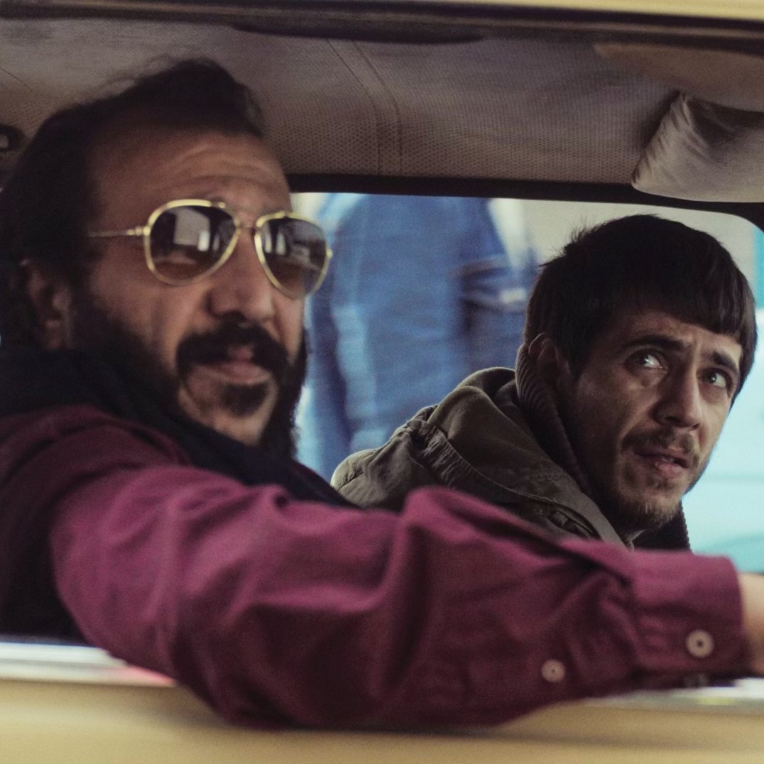 Deux hommes dans une voiture, l'un avec des lunettes de soleil, l'autre semble inquiet.