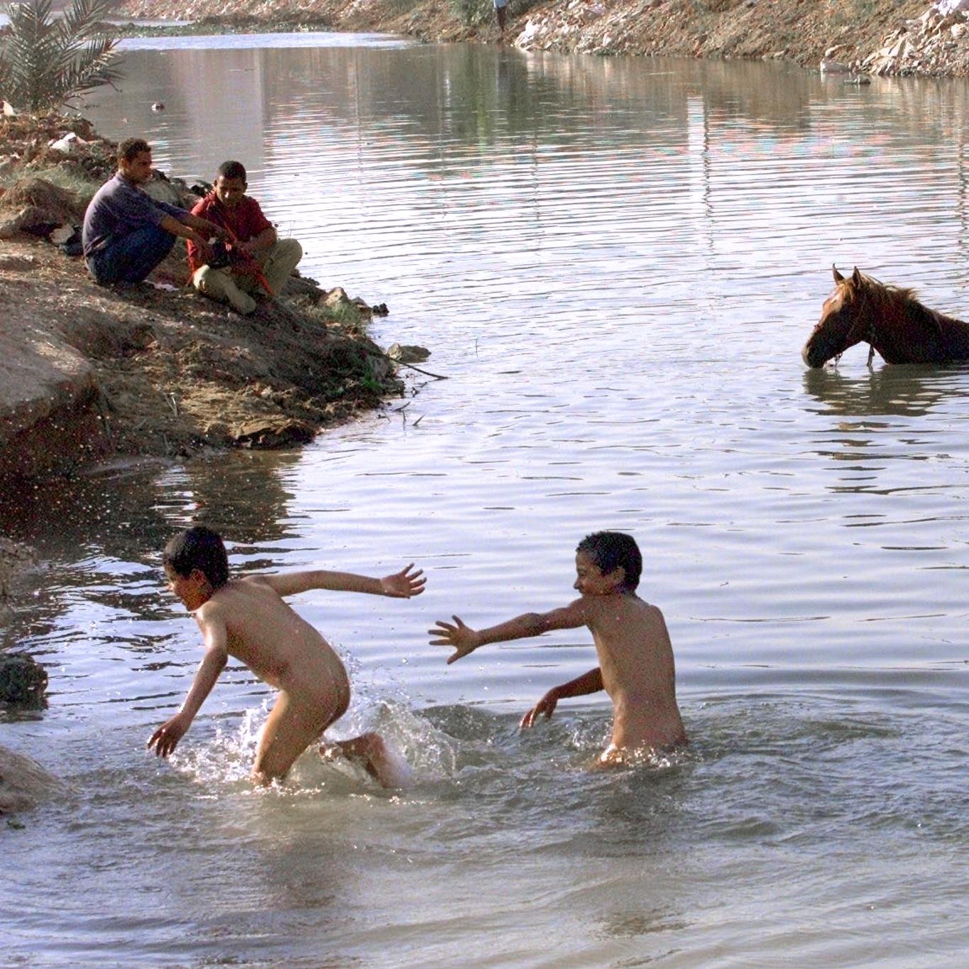 L'immagine mostra un fiume con due ragazzi che stanno giocando nell'acqua, apparentemente divertendosi a spruzzarsi. In lontananza, un cavallo si trova parzialmente immerso nell'acqua. Sulla riva, si notano anche alcune persone che osservano la scena. Gli elementi naturali e l'atmosfera sembrano suggerire un momento di spensieratezza e libertà durante una giornata calda.