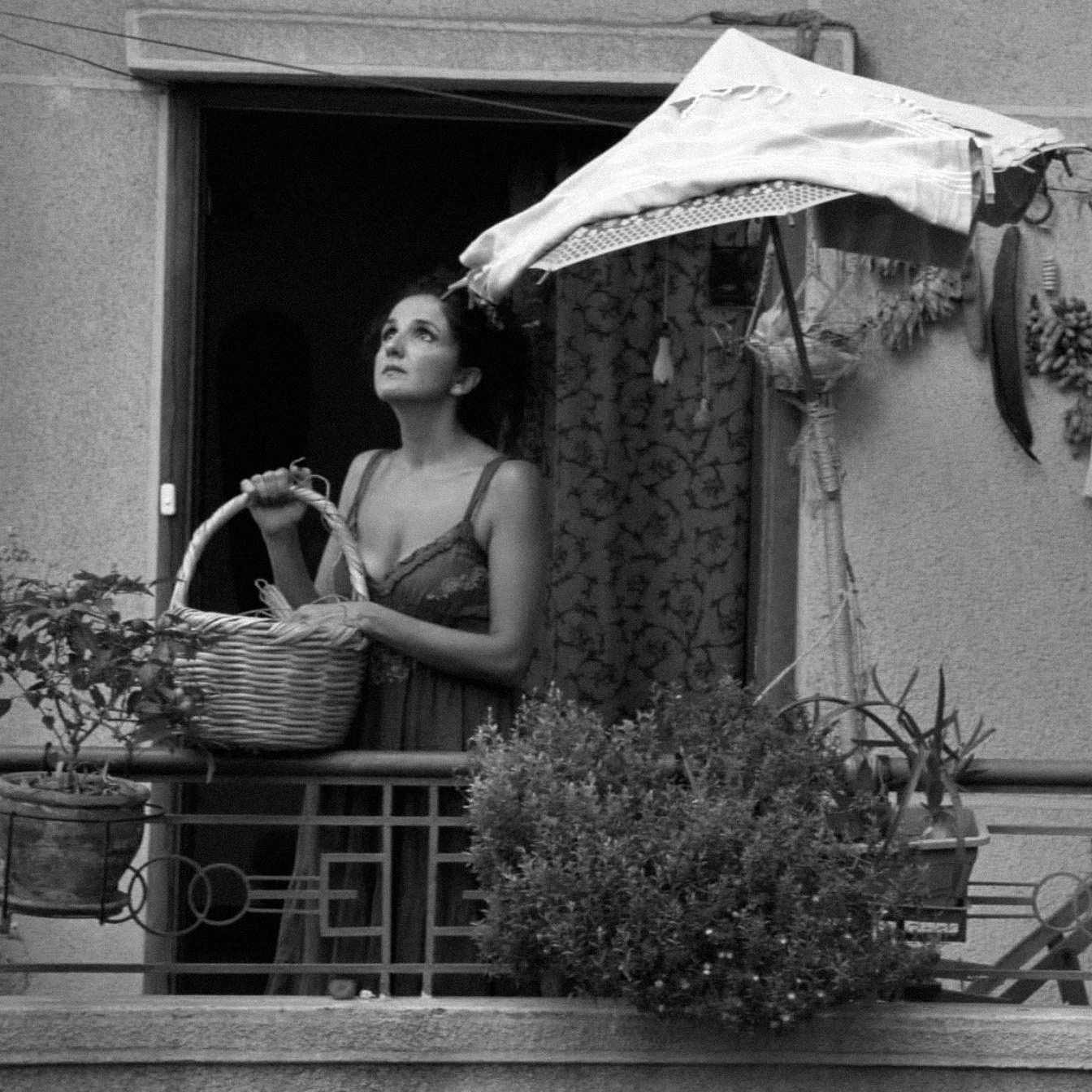 L'image montre une femme se tenant sur un balcon, tenant un panier dans ses bras. Elle semble contemplative, regardant au loin. Le balcon est décoré de plusieurs plantes et herbes suspendues. Un parasol en toile protège en partie la femme du soleil. L'image est en noir et blanc, ce qui lui donne une atmosphère nostalgique et mélancolique.
