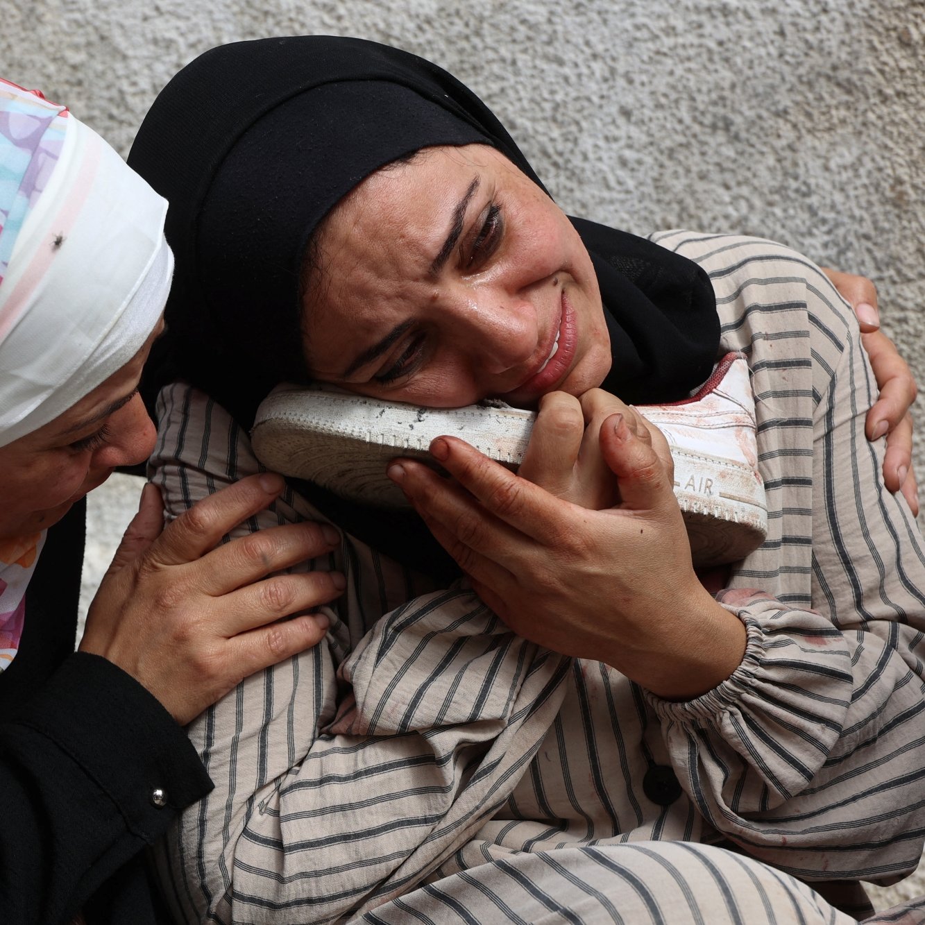 Two women in headscarves are embracing, expressing deep sorrow, with one holding a shoe.