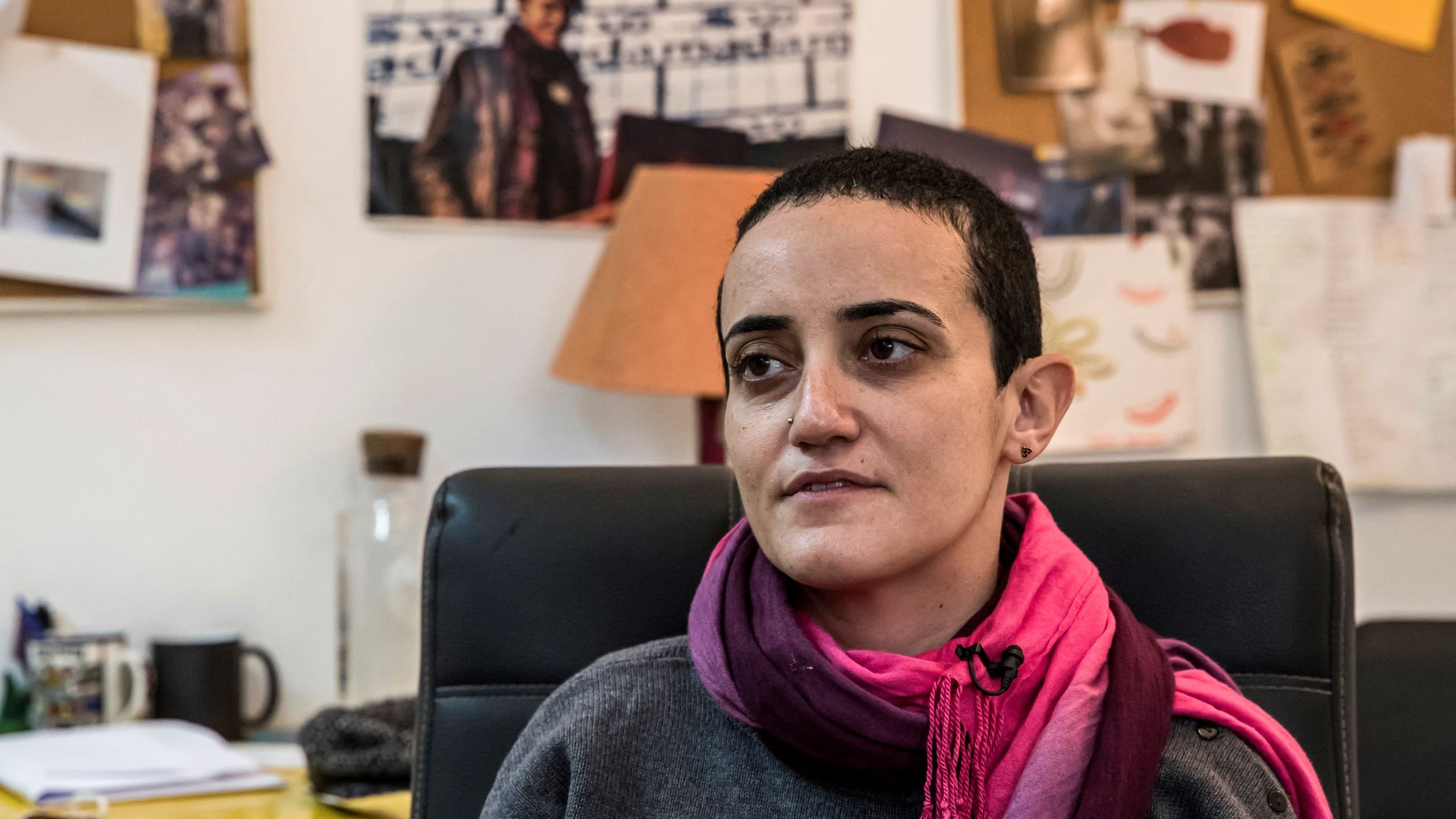 Je ne sais pas qui est présent sur cette image. Cependant, je peux te dire que la personne semble assise dans un bureau, portant un pull gris et une écharpe rose. On peut voir un environnement de travail avec des documents et des photos affichées en arrière-plan. L'éclairage donne une ambiance chaleureuse à la scène.