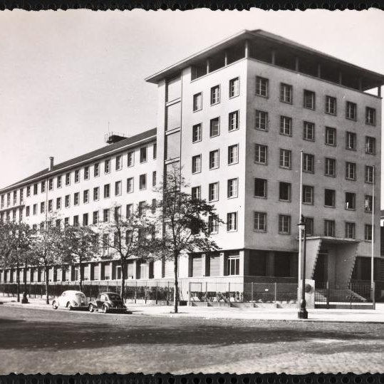 الصورة تُظهر مبنىً حديث التصميم، يتكون من عدة طوابق وله واجهة متنوعة الألوان. يتواجد حول المبنى مجموعة من الأشجار، ويبدو أن هناك شارعًا أمام المبنى حيث توجد بعض السيارات القديمة. يعكس تصميم المبنى طابعًا معماريًا بسيطًا وعصريًا، مما يوحي بأنه تم بنائه في فترة زمنية معينة في القرن العشرين. السماء تبدو واضحة مما يعطي انطباعًا بأن الصورة قد تم التقاطها في يوم مشمس.