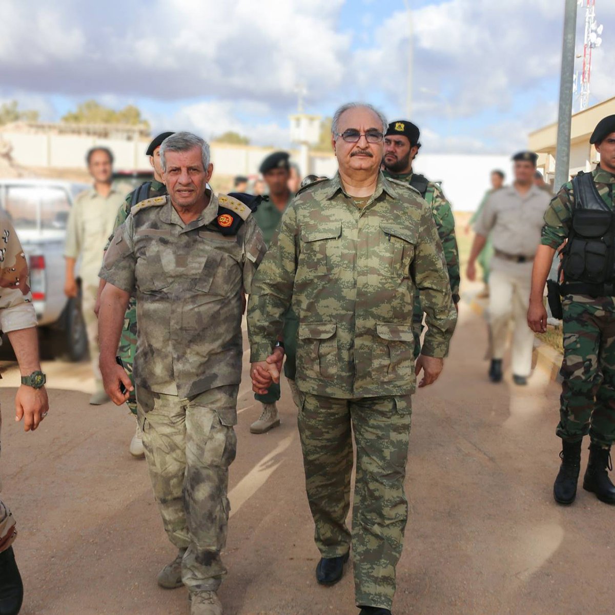 L'image montre un groupe de militaires marchant ensemble dans un environnement urbain. Deux hommes en avant-plan semblent discuter tout en marchant côte à côte. L'un d'eux porte un uniforme camouflage de couleur verte, tandis que l'autre a un uniforme militaire gris avec des distinctions. En arrière-plan, on peut voir d'autres soldats et des véhicules militaires, ce qui indique qu'ils se trouvent dans une zone potentiellement sécurisée. Les soldats semblent concentrés et en mission.