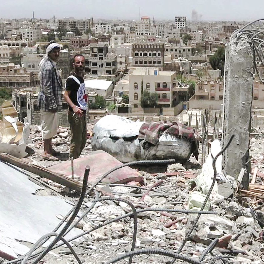 تظهر الصورة مشهدًا من مدينة مدمرة، حيث يقف رجلان على أنقاض مبنى. خلفهما، ترتفع مجموعة من المباني في المدينة، وهي تبدو متضررة بشكل كبير. يبدو أن البيئة المحيطة تعكس آثار الدمار، مع الحطام والمخلفات المنتشرة في كل مكان. السماء تبدو مشمسة، مما يبرز الف contrast بين الدمار والبيئة المحيطة.