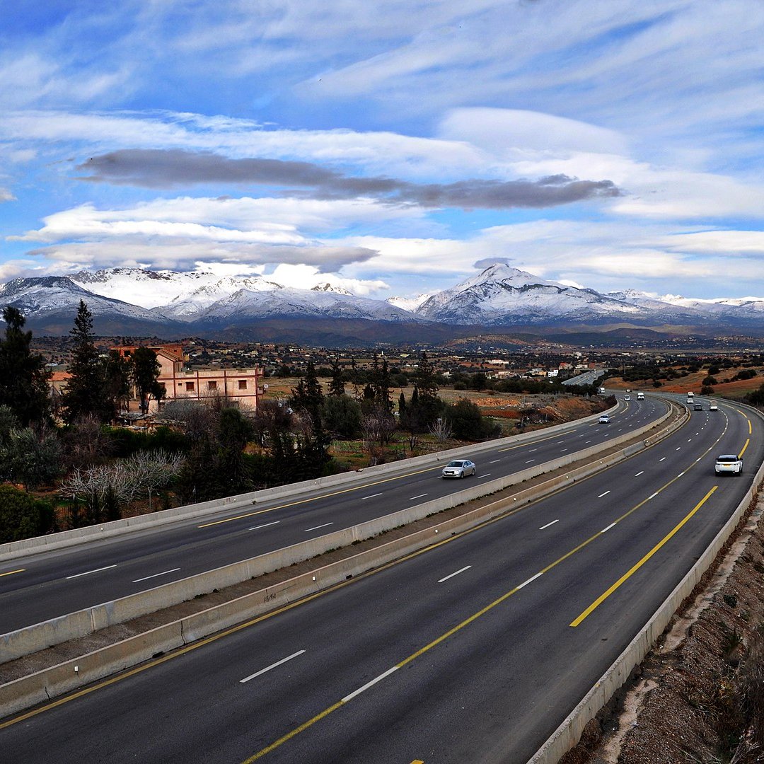 L'image montre une route large et bien entretenue, qui s'étend à travers un paysage pittoresque. On peut voir des montagnes majestueuses au loin, avec des sommets enneigés qui contrastent avec le ciel bleu parsemé de nuages. De chaque côté de la route, il y a quelques bâtiments et de la végétation, ce qui ajoute à la beauté de la scène. Cette vue évoque une ambiance paisible et naturelle, avec un mélange de modernité et de paysages ruraux.