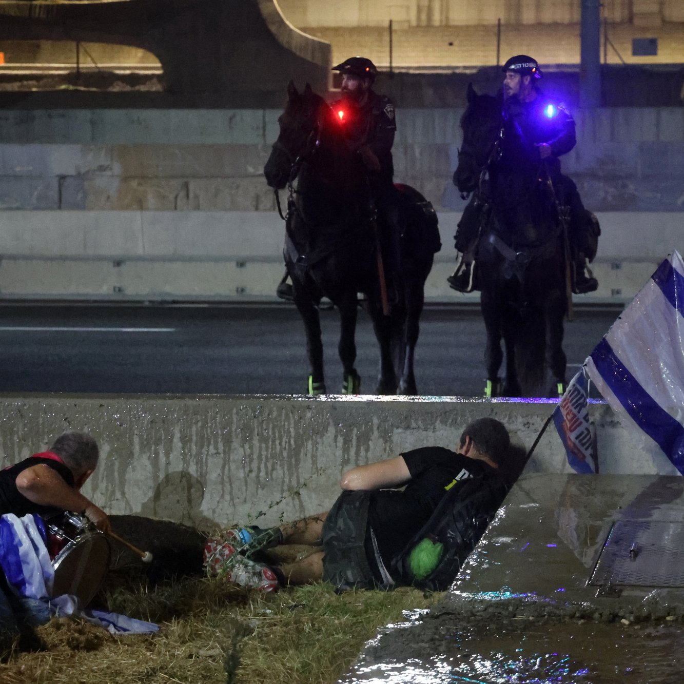 L'image montre une scène de tension en milieu urbain, avec deux policiers à cheval qui surveillent la situation. En bas, on aperçoit des manifestants, dont certains semblent se protéger ou être en détresse. Un drapeau israélien est visible au sol, ajoutant une dimension politique à la scène. L'éclairage des policiers et l'atmosphère générale suggèrent un climat de conflit ou de protestation.