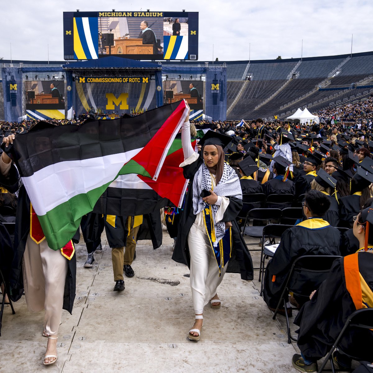 L'image montre une cérémonie de remise de diplômes qui se déroule dans un grand stade, probablement le Michigan Stadium. Des diplômés portant des toges et des chapeaux de cérémonie sont assis en rangées. Au premier plan, deux personnes tiennent des drapeaux : l'un est le drapeau palestinien et l'autre semble être un drapeau avec des couleurs distinctives. À l'arrière-plan, un grand écran affiche un orateur. L'ambiance est festive et solennelle, typique des cérémonies de graduation.