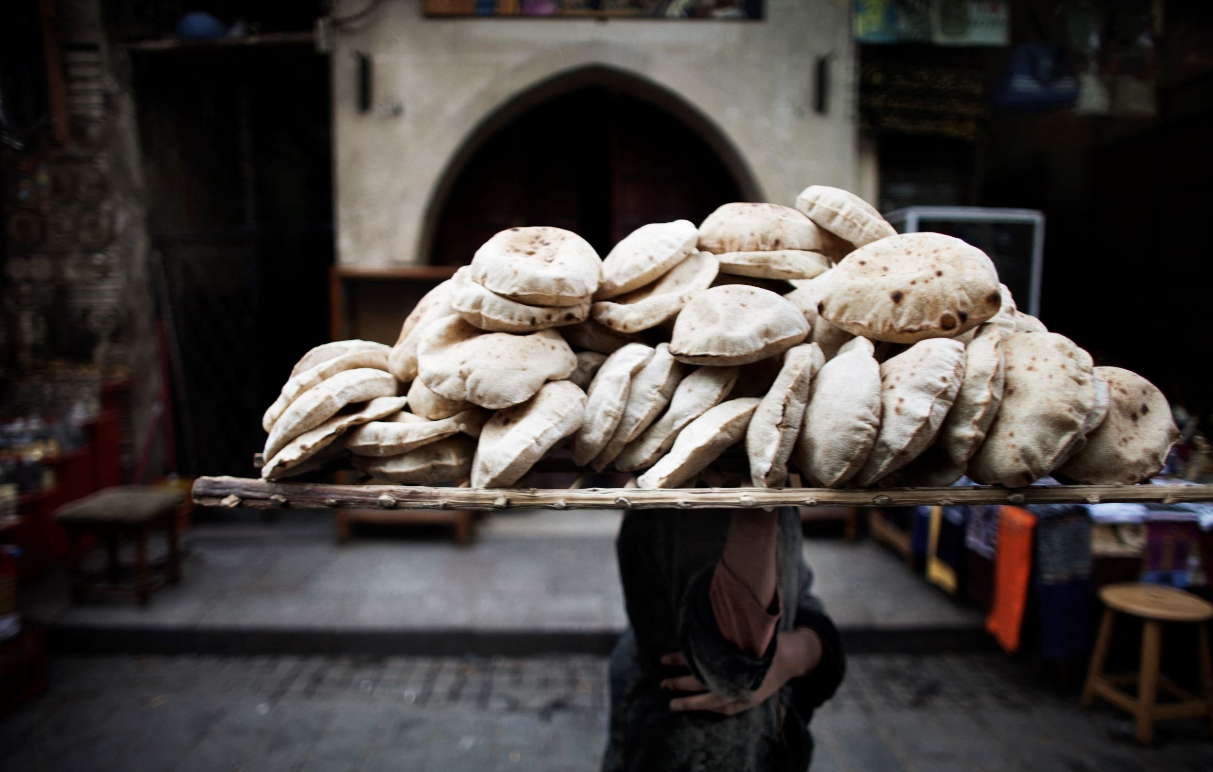 L'image montre une personne portant une grande pile de pains sur un plateau placé sur sa tête. Les pains, ronds et légèrement plats, semblent fraîchement cuits et sont empilés de manière ordonnée. En arrière-plan, on peut apercevoir des éléments typiques d'un marché, avec des structures architecturales anciennes et un éclairage ambiant qui donne une atmosphère vivante à la scène. L'accent est mis sur le transport traditionnel des marchandises en milieu urbain.