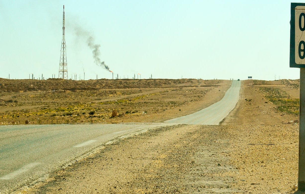 L'image montre un paysage désertique avec une route qui s'étend à perte de vue. À droite, un panneau indiquant "Hassi R'Mel" avec des chiffres. En arrière-plan, on aperçoit une tour de communication et une colonne de fumée émanant d'une installation, probablement liée à l'industrie pétrolière. Le sol est aride, et il n'y a pas beaucoup de végétation environnante, illustrant un environnement rural et isolé.