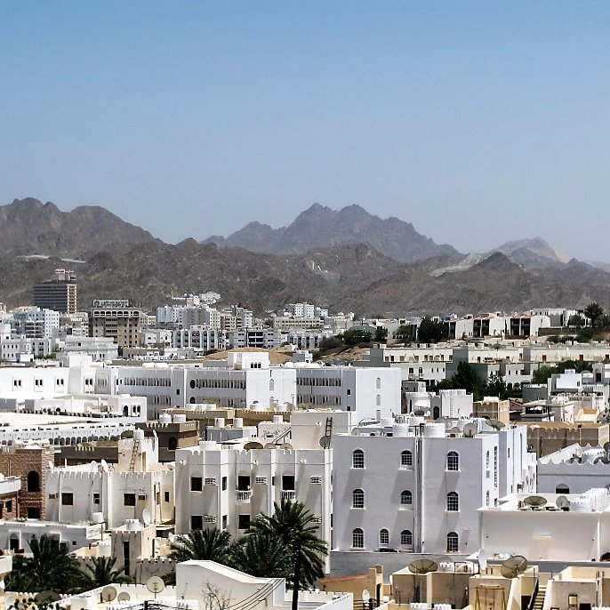 L'image montre une vue panoramique d'une ville située dans une région montagneuse. On peut voir de nombreux bâtiments aux façades blanches et crème, qui typiquement se distinguent dans un environnement aride. Les montagnes en arrière-plan ajoutent une dimension naturelle impressionnante au paysage urbain. Le ciel est dégagé et ensoleillé, accentuant la lumière qui se reflète sur les bâtiments. Cette scène évoque une atmosphère calme et sereine.