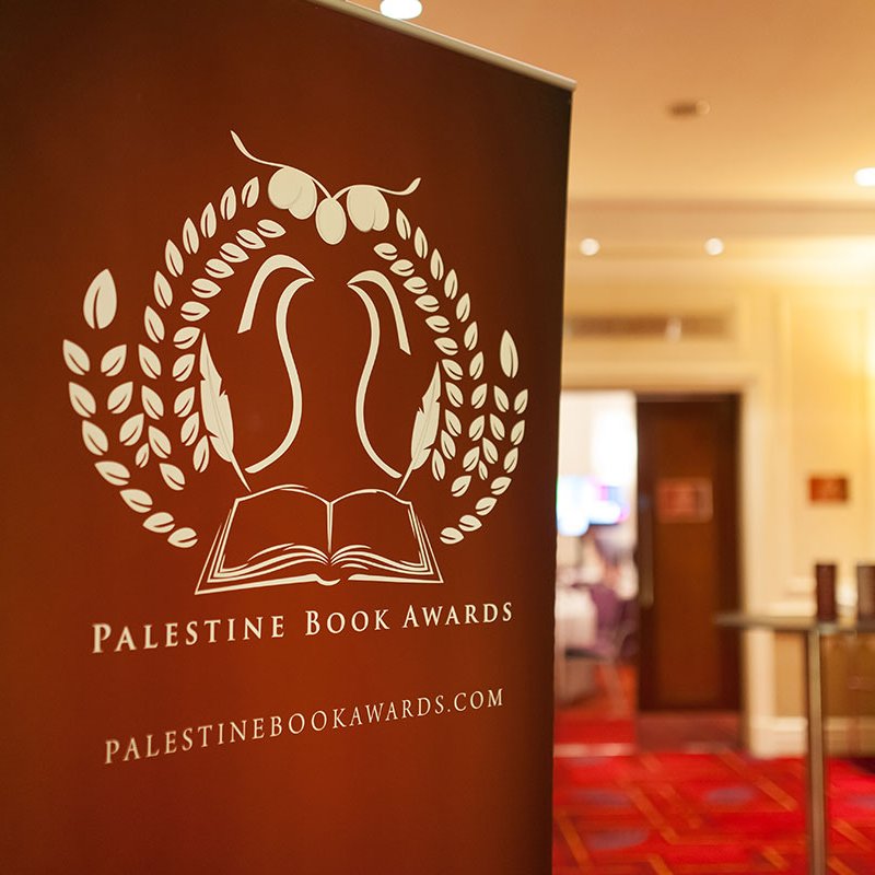 L'image représente un panneau de présentation pour les Palestine Book Awards. Ce panneau arbore un design avec des éléments graphiques, incluant une illustration d'un livre ouvert entouré de feuilles. Le texte indique "Palestine Book Awards" ainsi que le site web associé. L'arrière-plan semble être une salle de réception, avec un sol tapissé et des éclairages doux.