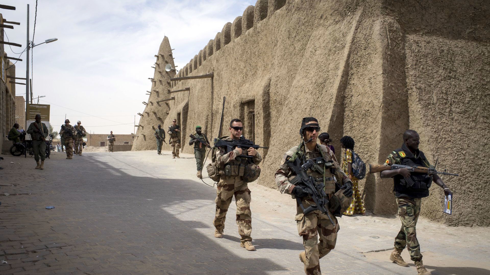 La imagen muestra a un grupo de soldados que caminan por una calle en una zona con arquitectura de barro. A su alrededor, se pueden ver estructuras típicas del lugar, como muros de adobe. Los soldados llevan equipos militares y armamento, y algunos parecen estar en formación o realizando patrullas. La escena sugiere un ambiente de seguridad o una operación militar en un contexto urbano o semiurbano.