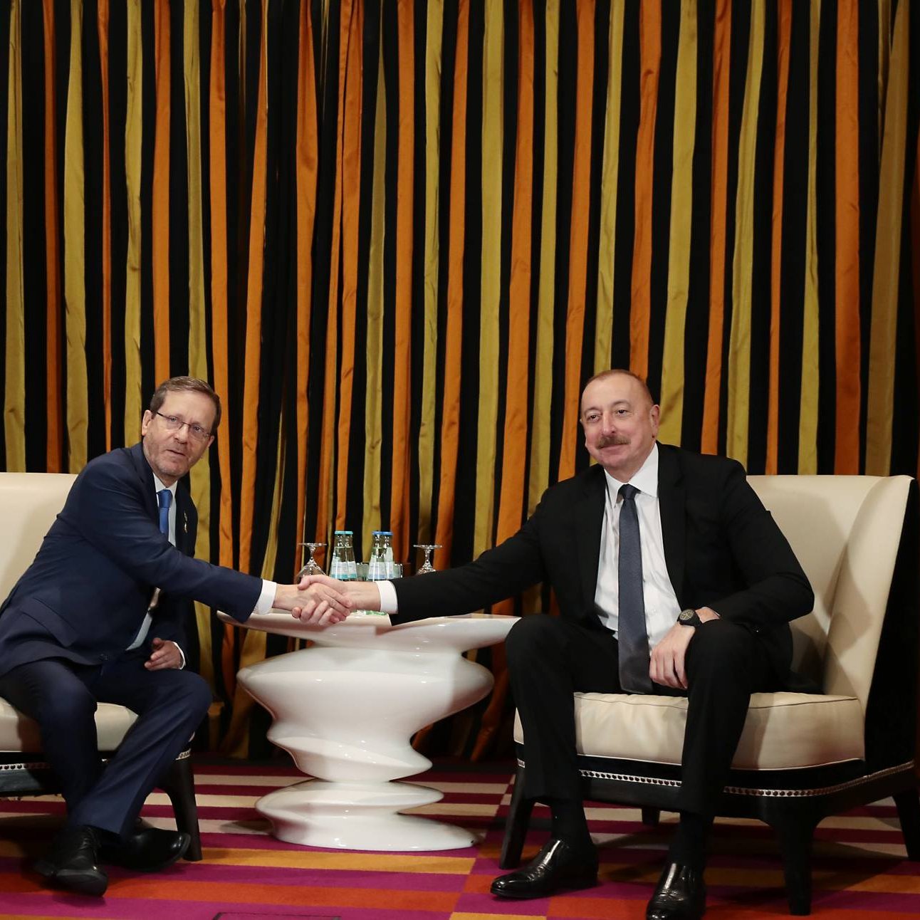 دو مرد در حال دست دادن نشسته‌اند. پرچم‌های اسرائیل و آذربایجان در پس‌زمینه هستند. رنگ‌ها گرم و زنده‌اند.