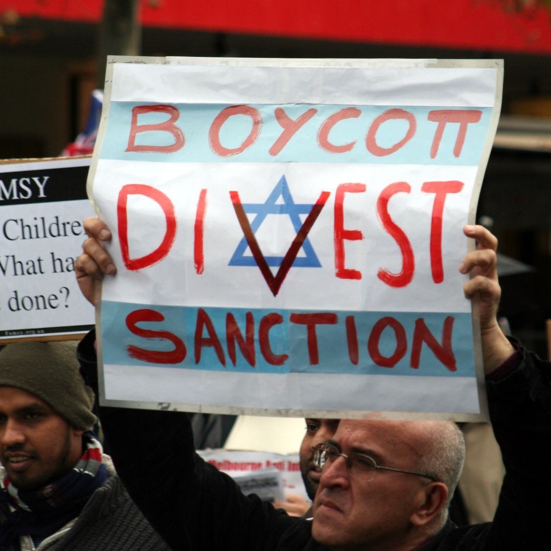 L'image montre des manifestants tenant des pancartes lors d'une manifestation. L'une des pancartes, bien visible, porte les mots "BOYCOTT, DIVEST, SANCTION" avec un symbole en forme d'étoile. Les manifestants semblent exprimés des opinions politiques ou sociales fortes, et l'ambiance de l'événement semble engagée.