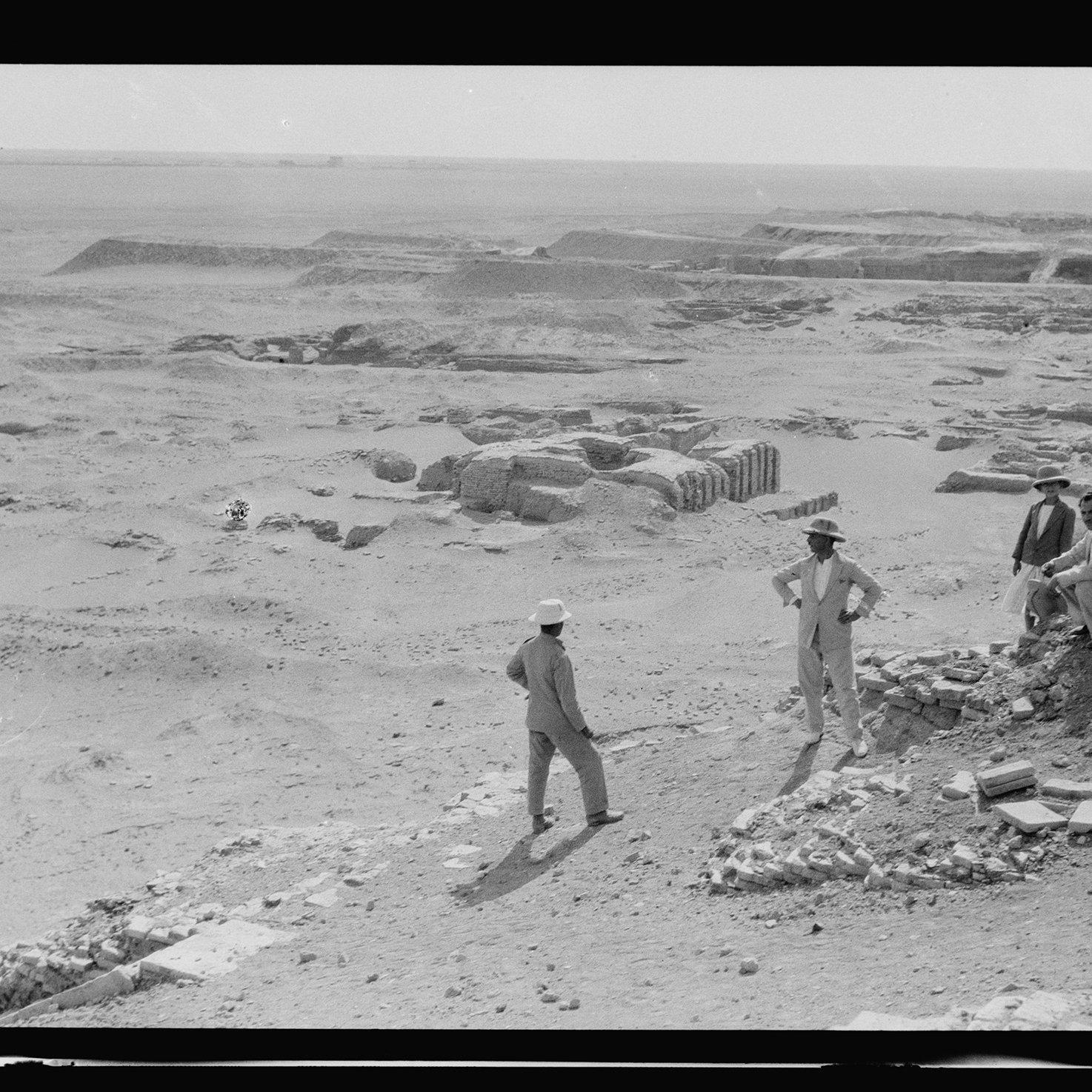 L'image montre une scène archéologique dans un paysage désertique. Plusieurs hommes, habillés de manière décontractée et portant des chapeaux, sont visibles. Ils semblent discuter ou examiner des fragments de pierres et des structures en ruines au sol. En arrière-plan, on peut voir des sites de fouille avec des vestiges qui suggèrent une activité archéologique en cours. L'environnement est aride, et le ciel paraît clair, évoquant une ambiance de travail sur un site ancien.